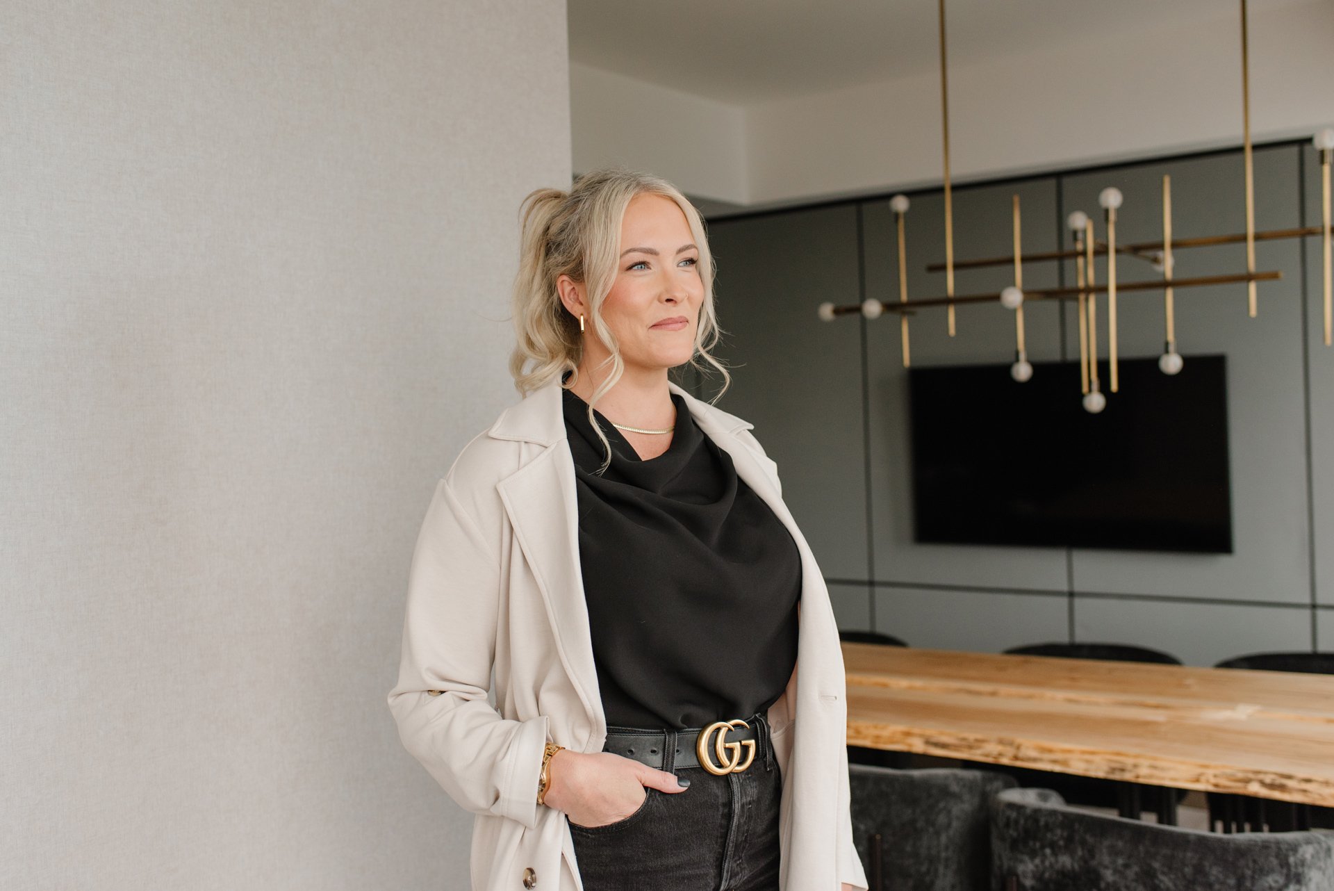 Jenna Shaw wearing a white coat over a black top, standing in a modern indoor space with a large wood table and a geometric chandelier in the background.