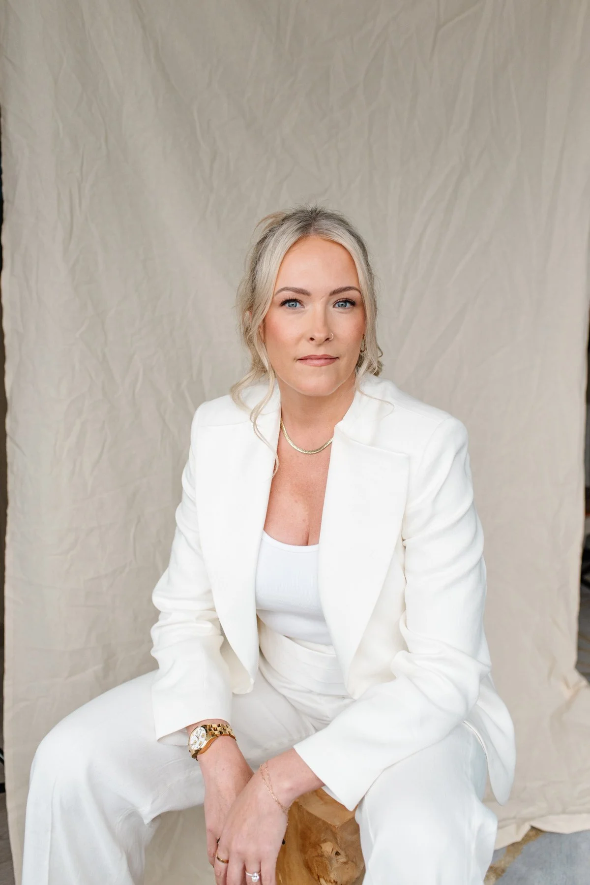 Jenna Shaw seated in a white suit and gold watch, looking at the camera against a linen backdrop