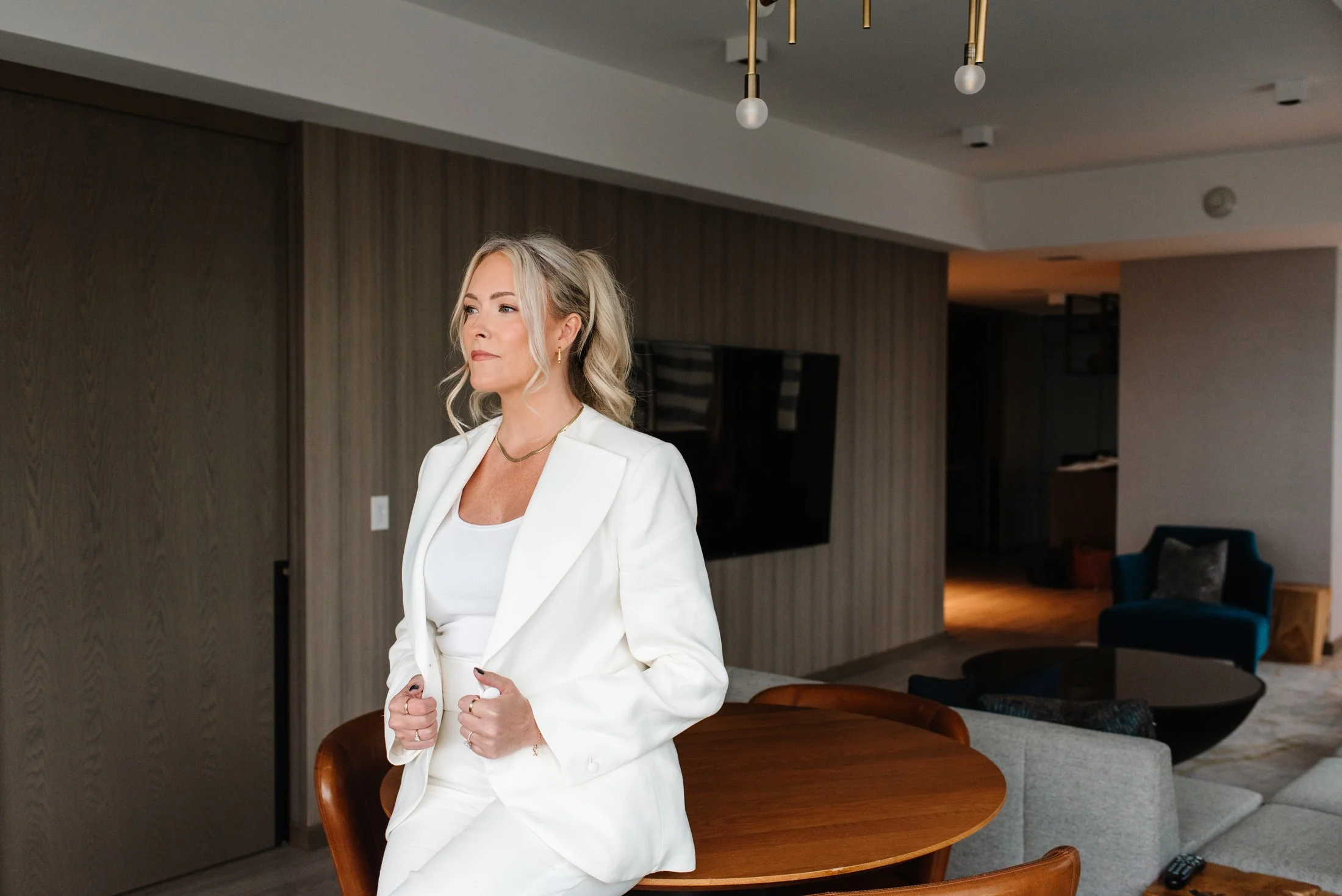 Jenna Shaw in a white suit leaning against a walnut table in a modern interior, looking to the side