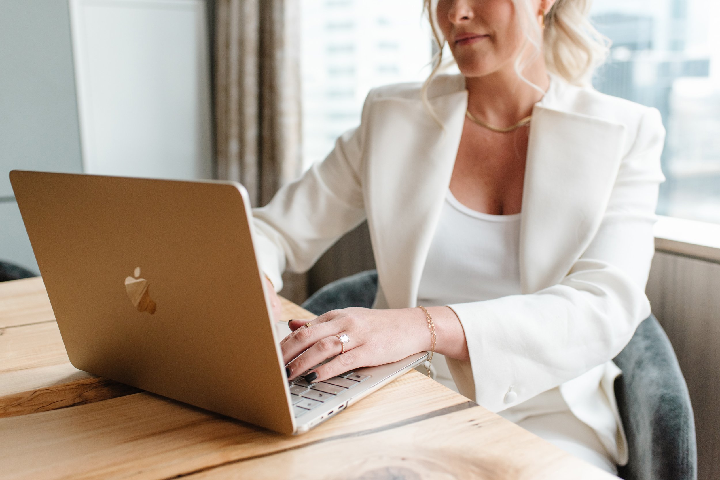 Close-up of hands typing on a laptop at a wooden table, wearing a white blazer and gold jewellery