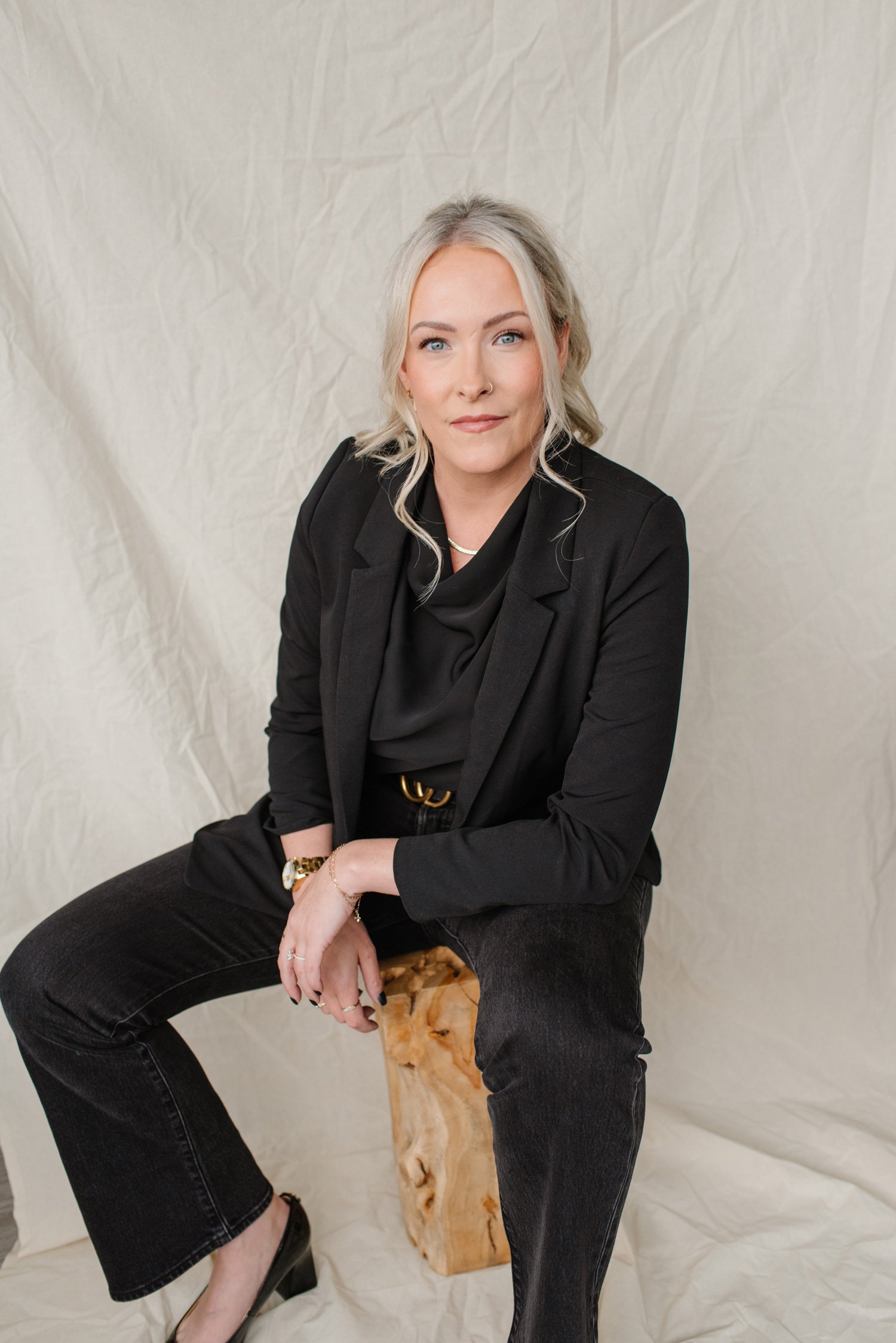 Jenna Shaw seated on a wooden stool in a black blazer and jeans against a linen backdrop