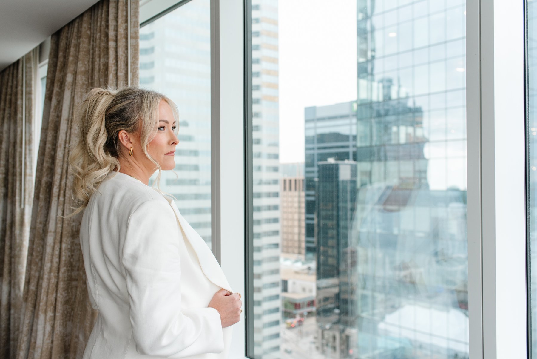 Jenna Shaw in a white blazer looking out a high-rise window over the downtown skyline