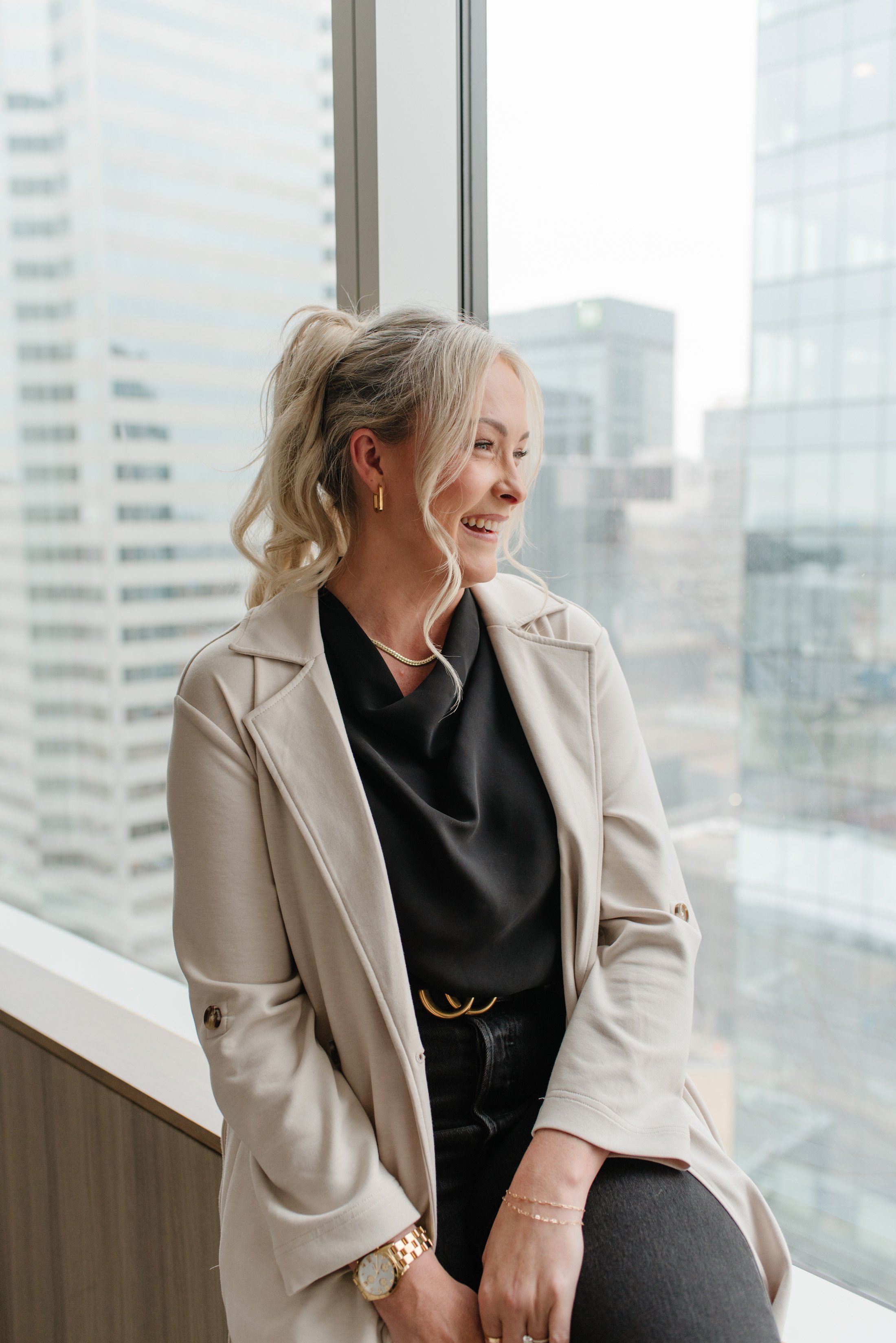 Jenna Shaw laughing beside a city-view window in a beige trench coat and black blouse