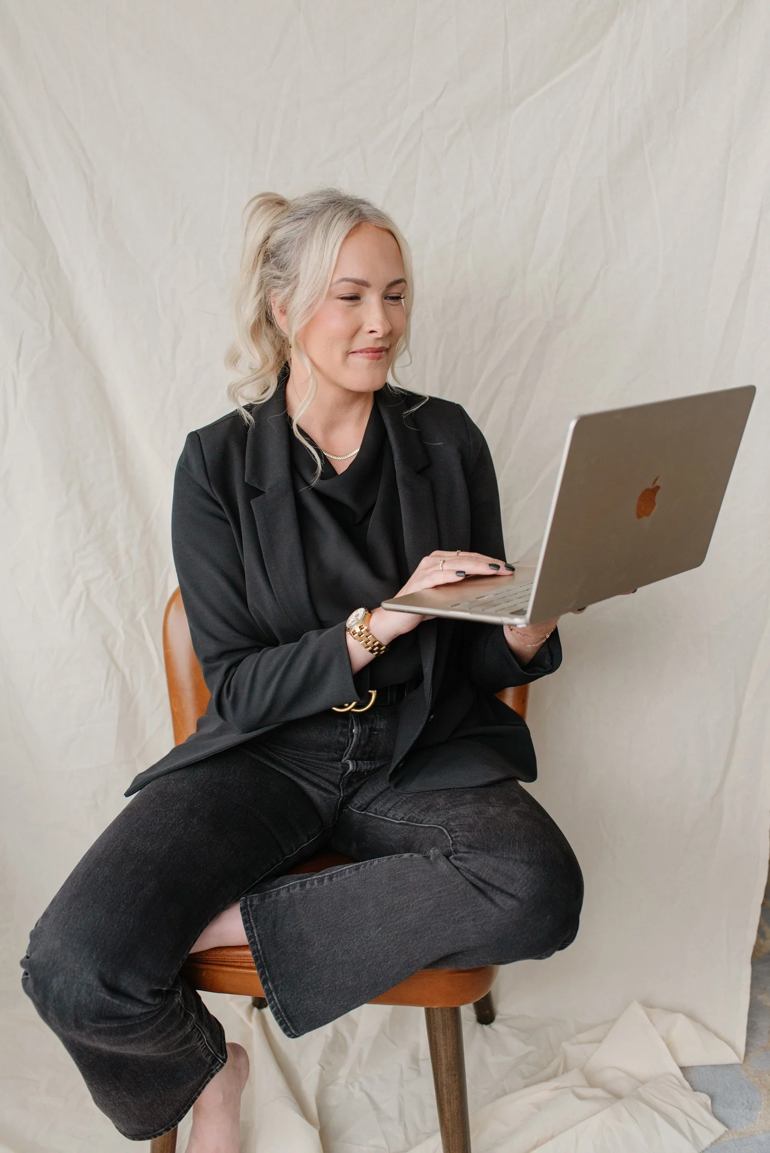 Jenna Shaw smiling at a laptop, seated cross-legged on a leather chair in a black blazer