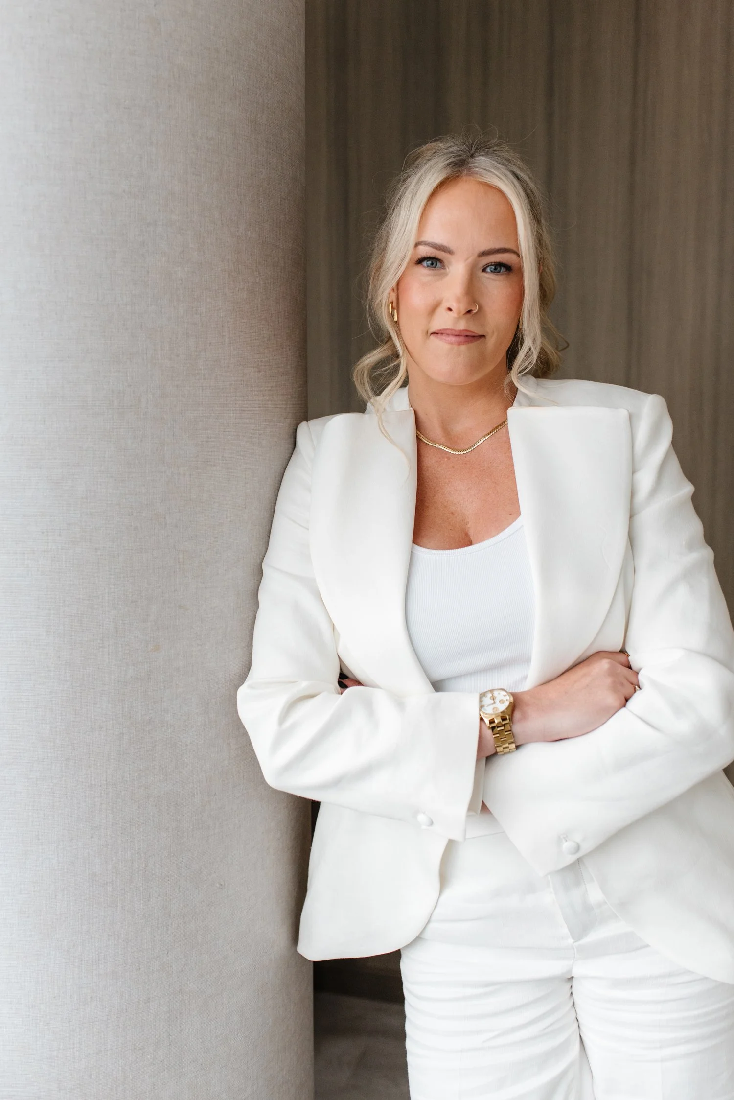 Jenna Shaw wearing a white blazer, white top, gold jewelry, and a gold watch, standing with her arms crossed and leaning against a beige wall.