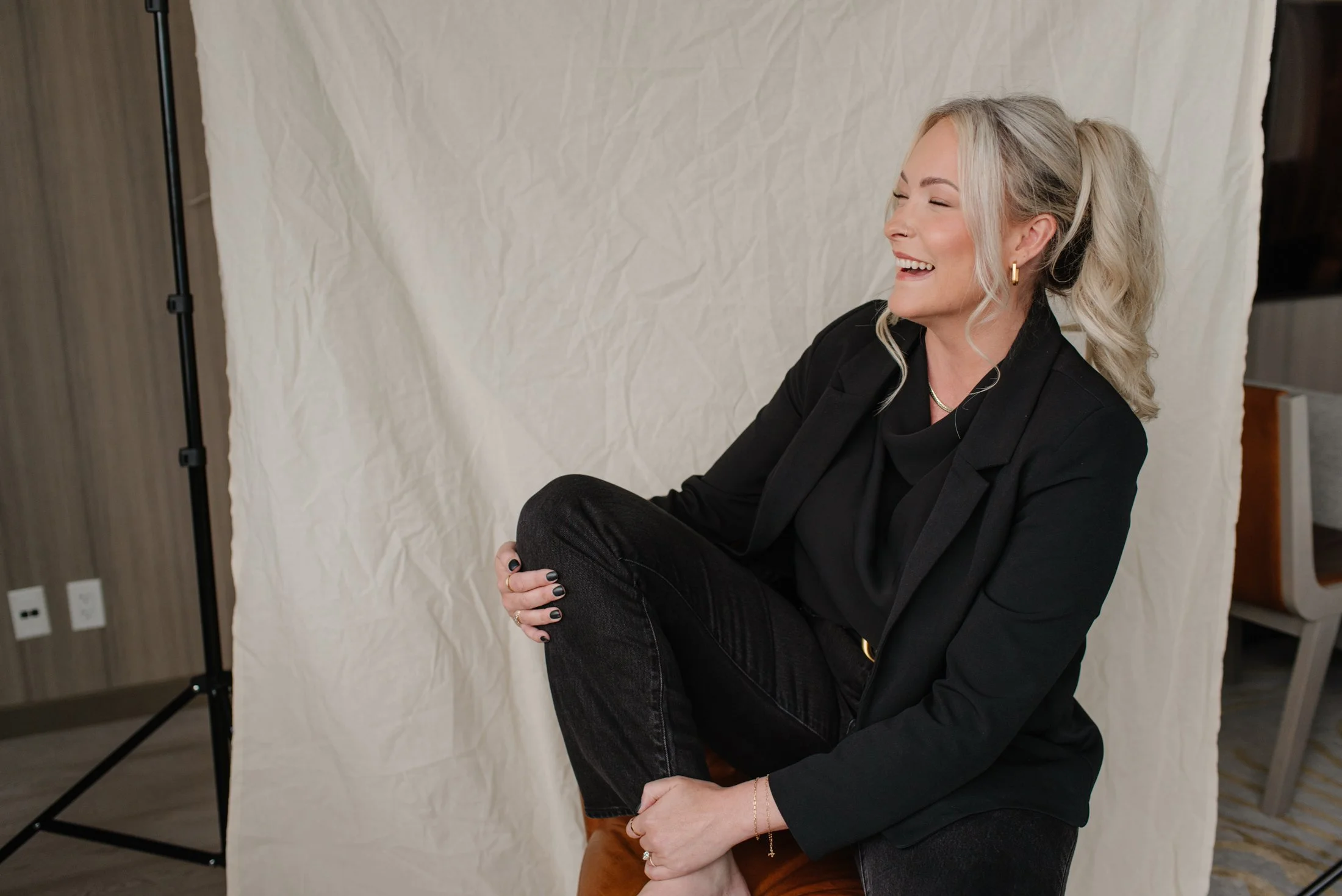Jenna Shaw laughing with one knee raised, seated in a black blazer during a portrait session