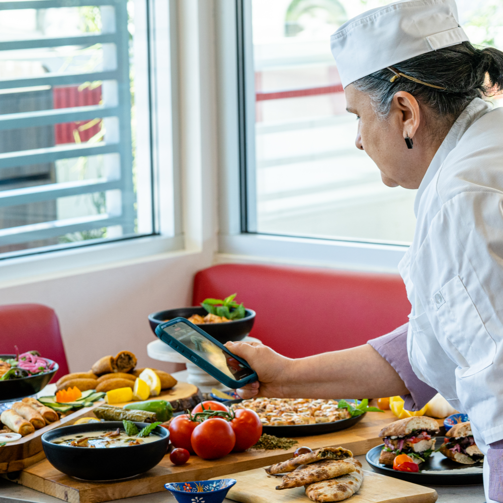 A chef taking a photo of a spread of various dishes on a table with fresh vegetables and baked goods, in a well-lit kitchen or dining area.