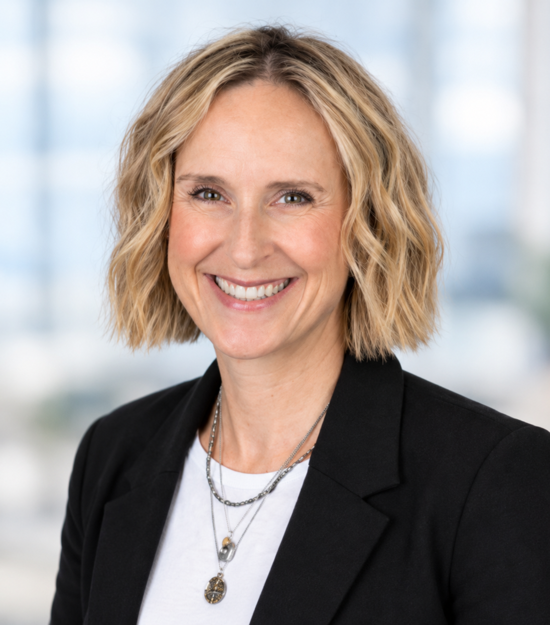 Portrait of a smiling woman with blonde, wavy hair wearing a black blazer and layered necklaces against a blurred office background.