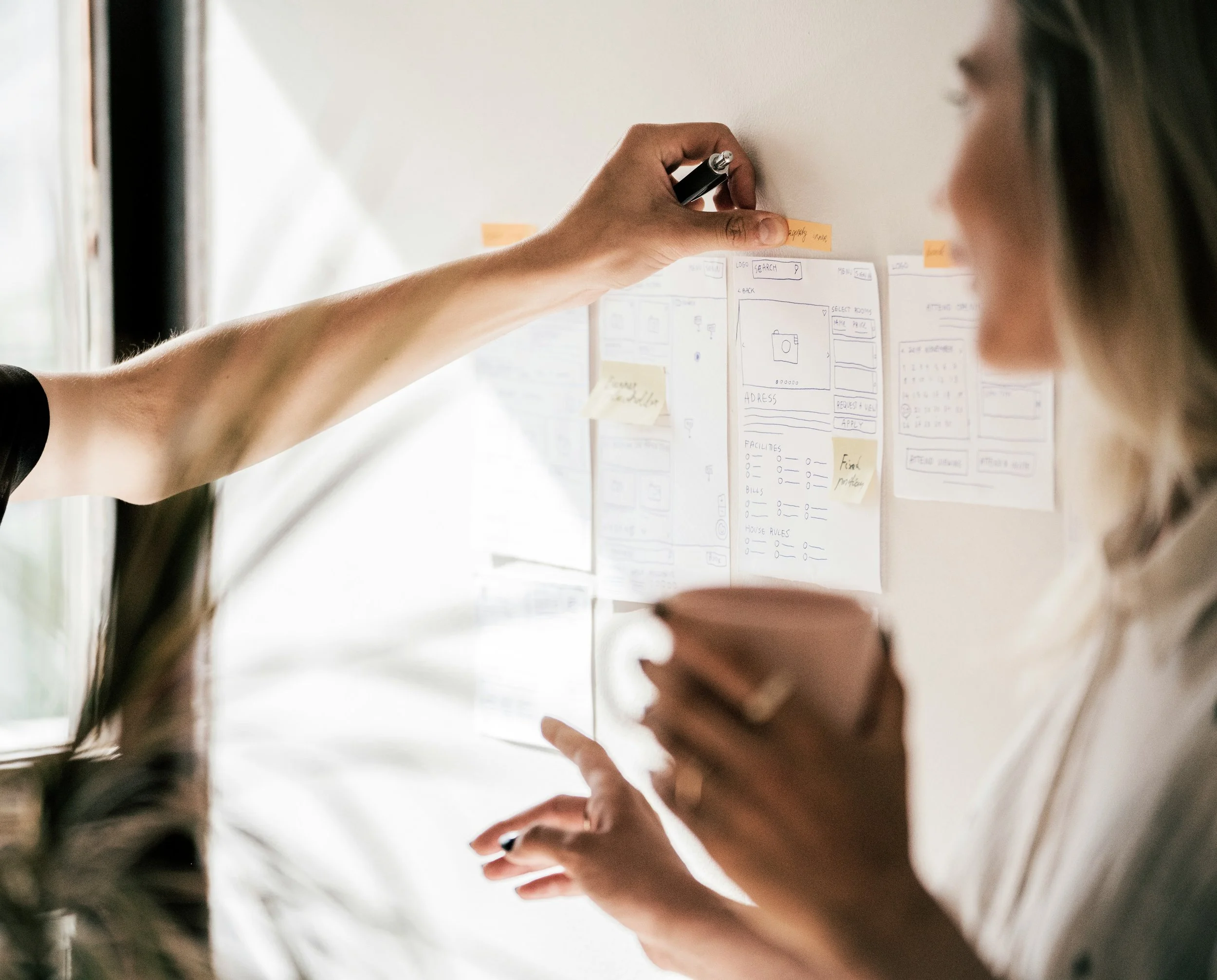 A person is placing sticky notes on a wall with notes and sketches, wearing glasses and holding a marker.