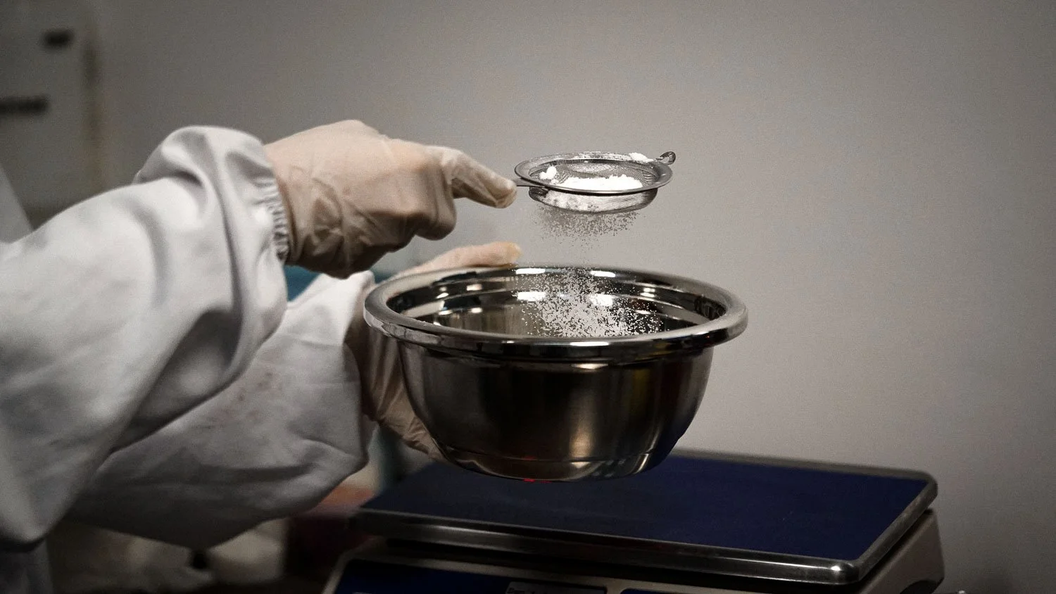 Person wearing gloves sifting powder through a metal sieve into a stainless steel bowl on a digital scale.