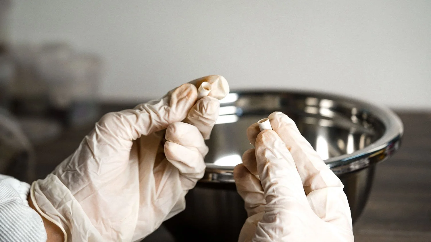 Hands wearing gloves holding a small, hollow, white capsule medicine, with a metal mixing bowl in the background.