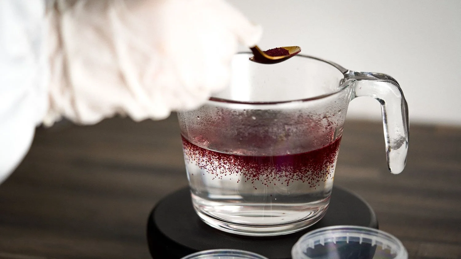 A glass cup with a handle filled with clear liquid and pinkish powder sprinkled around the rim, atop a black coaster on a gray wooden surface, with a white towel and small containers nearby.