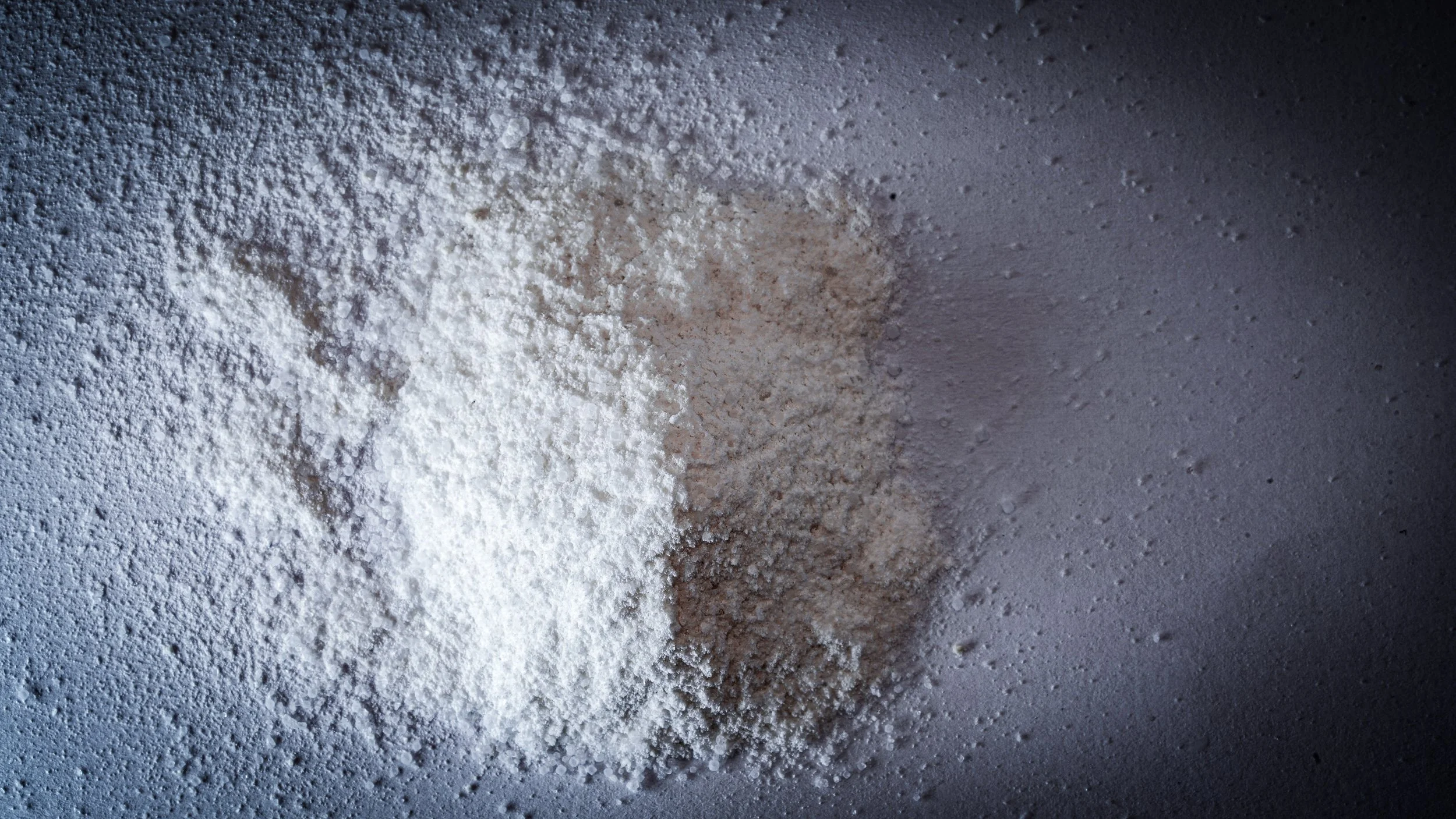 Close-up of a surface with white powder, possibly flour or powdered sugar, scattered unevenly on a flat surface.