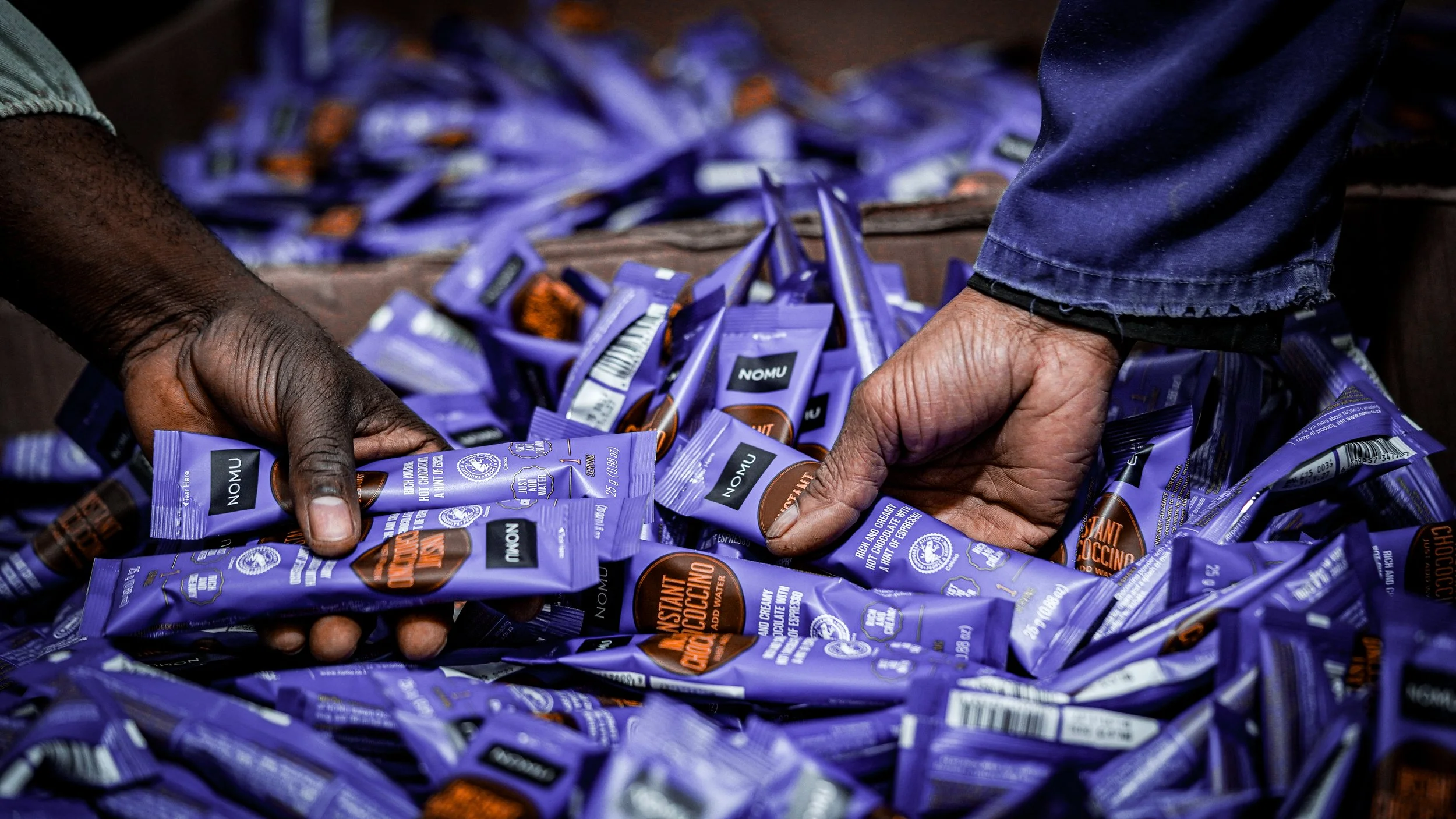 Hands arranging purple packages of Nomu instant coffee on a table.