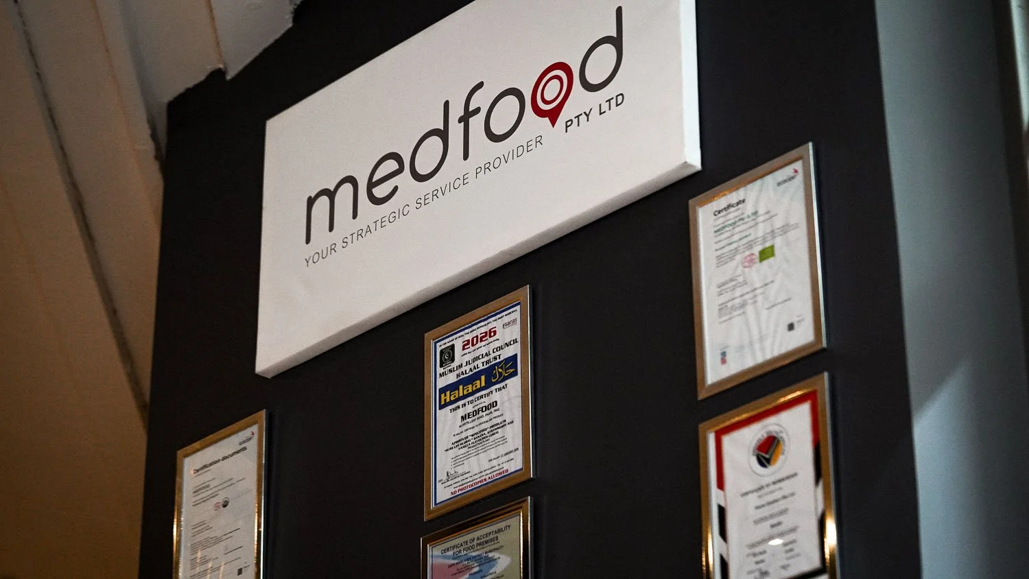 A black wall featuring a white sign with black and red text that reads "medfood your strategic service provider ptY LTD". Below the sign are multiple framed certificates and documents.