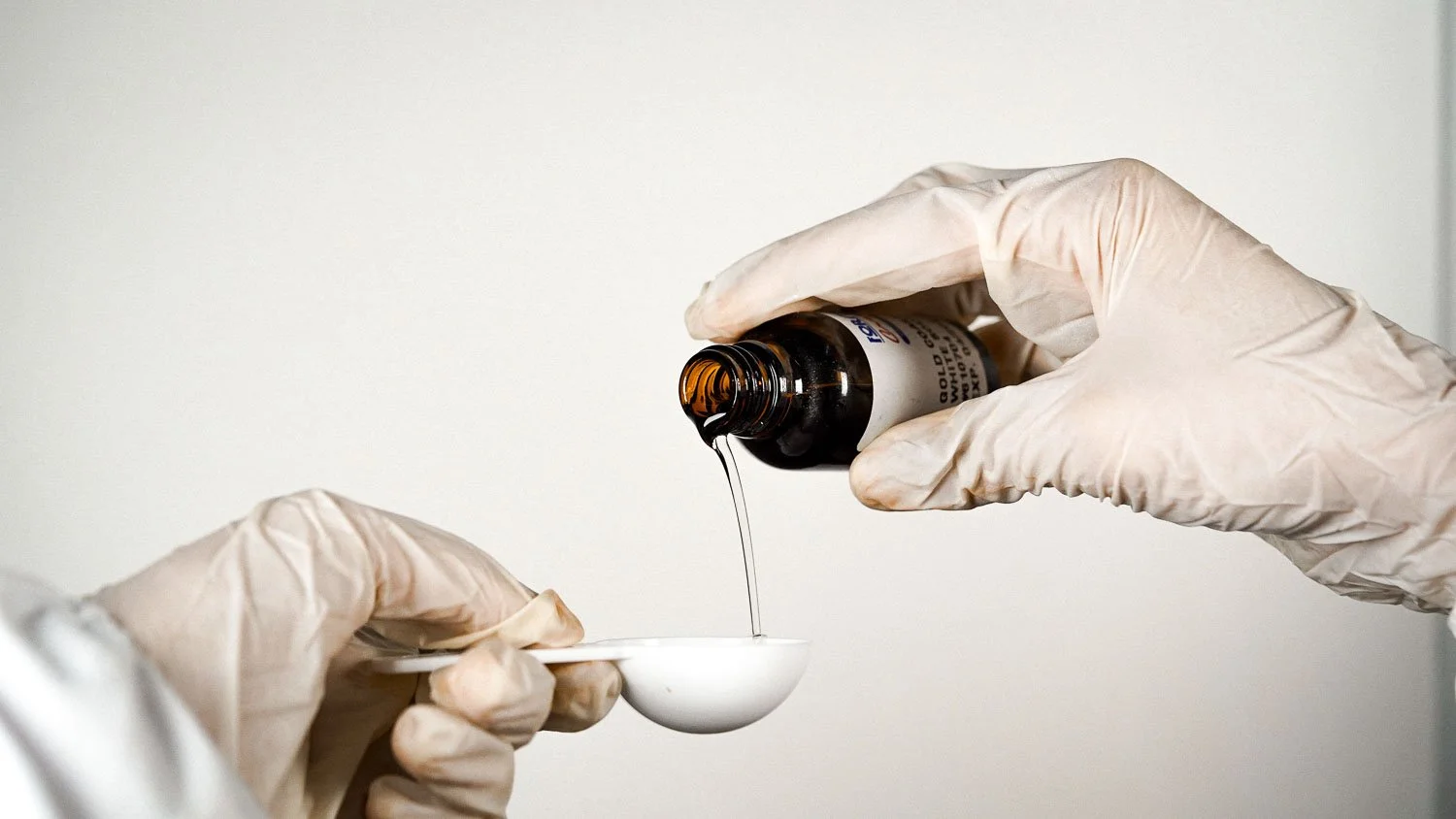 A person wearing medical gloves pouring a liquid from a brown medicine bottle into a white measuring spoon.