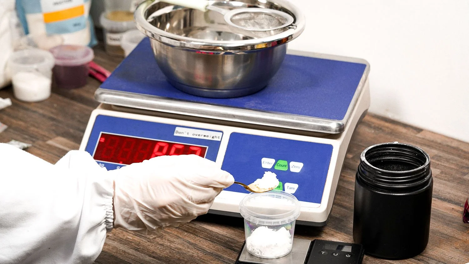 Person in white gloves weighing a white powdery substance on a digital scale in a laboratory or production setting with various containers and equipment around.