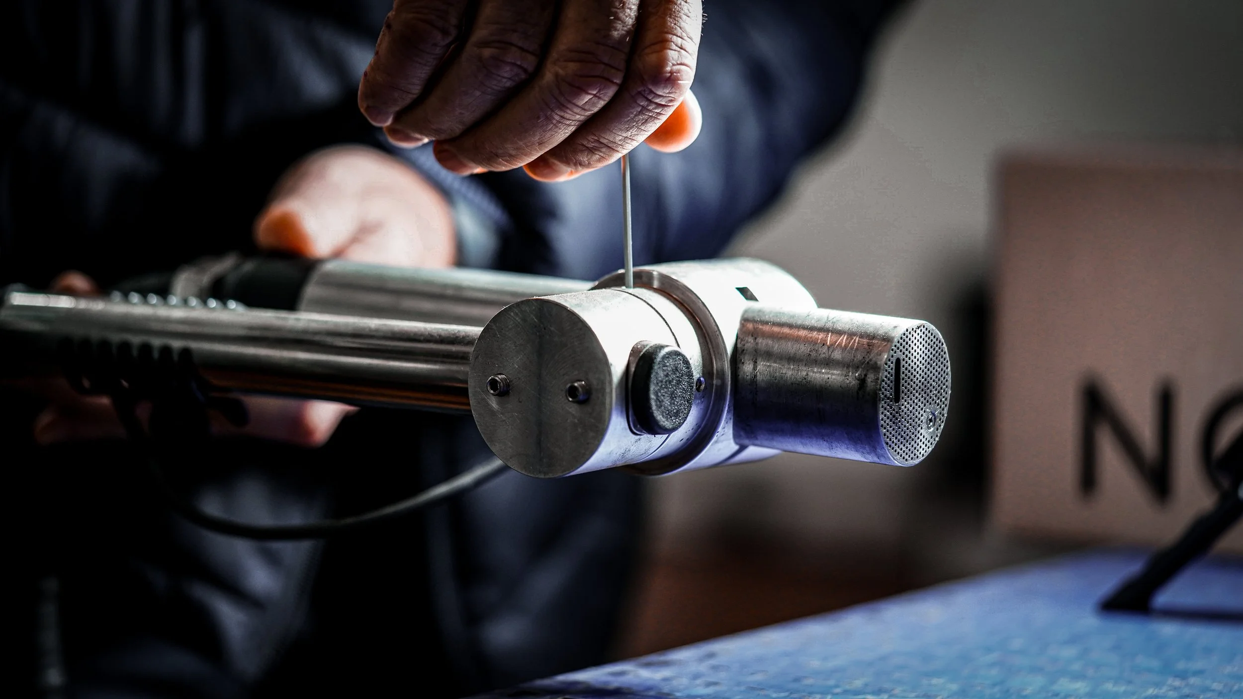 Close-up of a person’s hand using a tool on a metal cylindrical piece, with ergonomic grip and gear mechanisms visible, in a workshop or manufacturing setting.