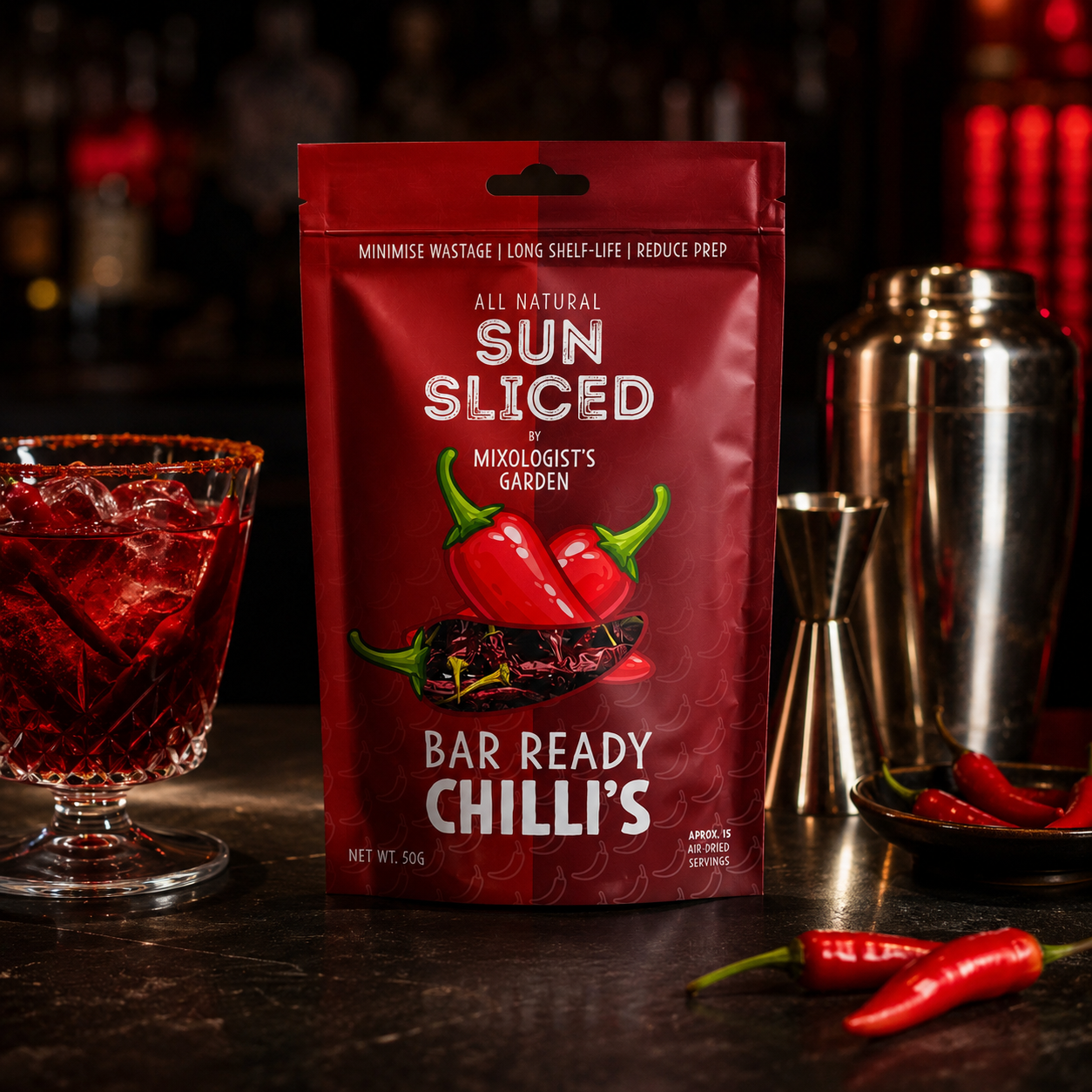 Package of all-natural sun-sliced chili peppers from Mixologist's Garden, bar-ready with chili illustration on the front, placed on a dark bar counter with a glass of red beverage, a metal cocktail shaker, and a bowl of fresh chili peppers nearby.