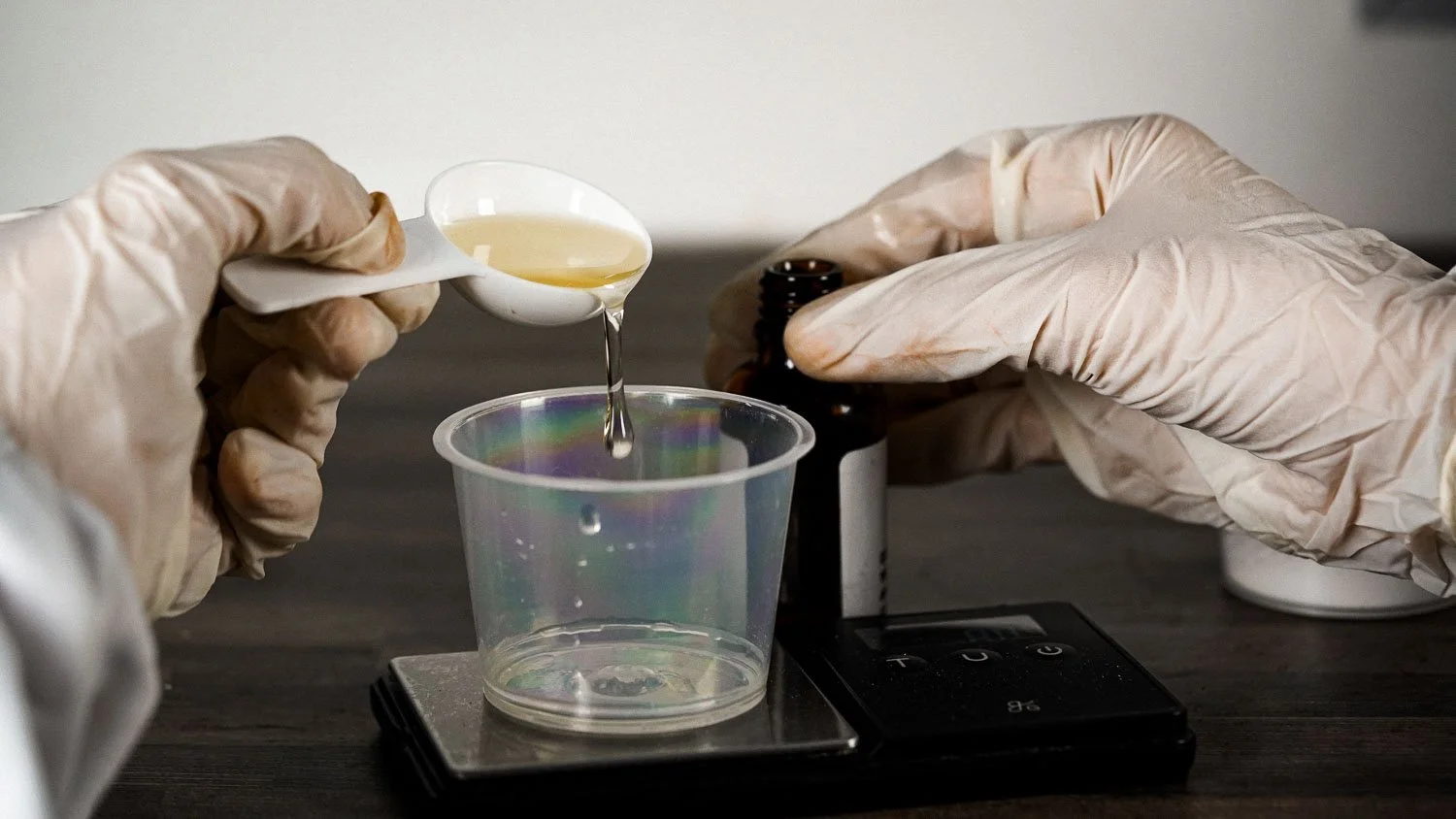 Hands wearing gloves measuring a yellow liquid with a white scoop above a scale, with a brown bottle in the background.