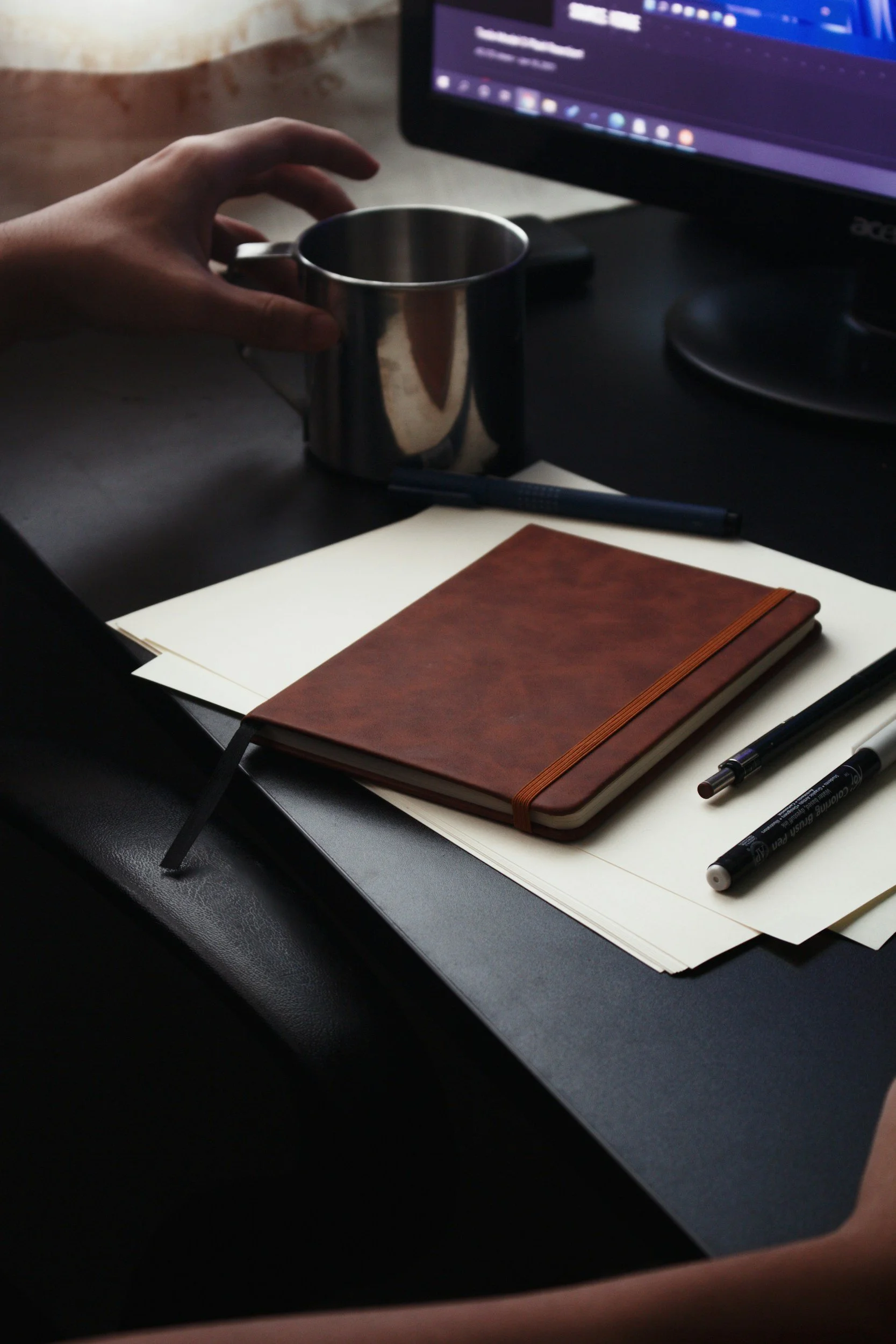 A workspace with a person holding a metallic coffee mug, a closed brown leather notebook, a black pen, and two black marker pens on a white sheet of paper, with a computer monitor displaying a dark themed interface in the background.