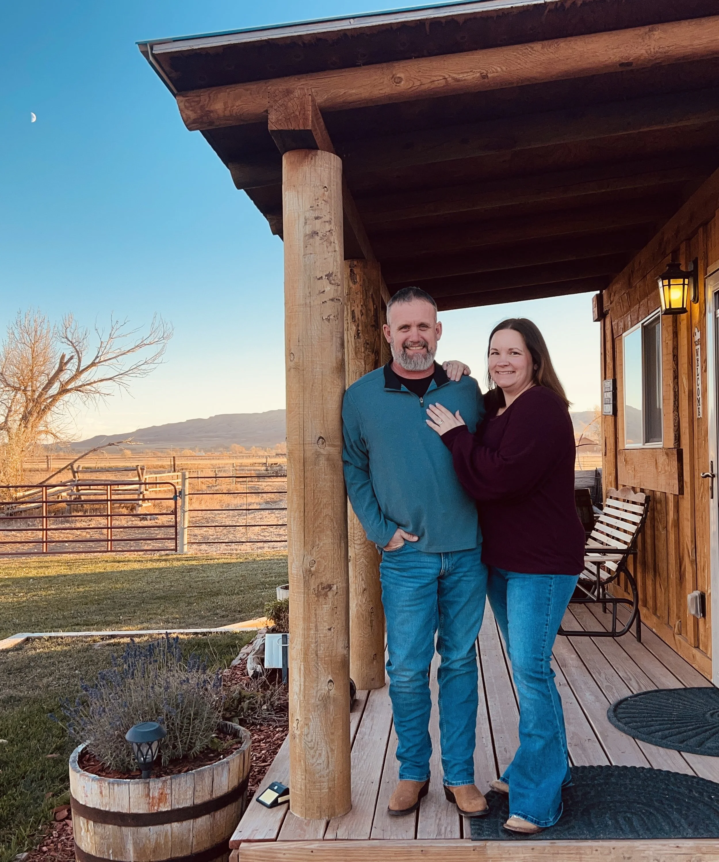 A couple standing on a wooden porch outdoors at sunset, smiling at the camera, with a mountain range in the background and a clear sky with the moon visible.