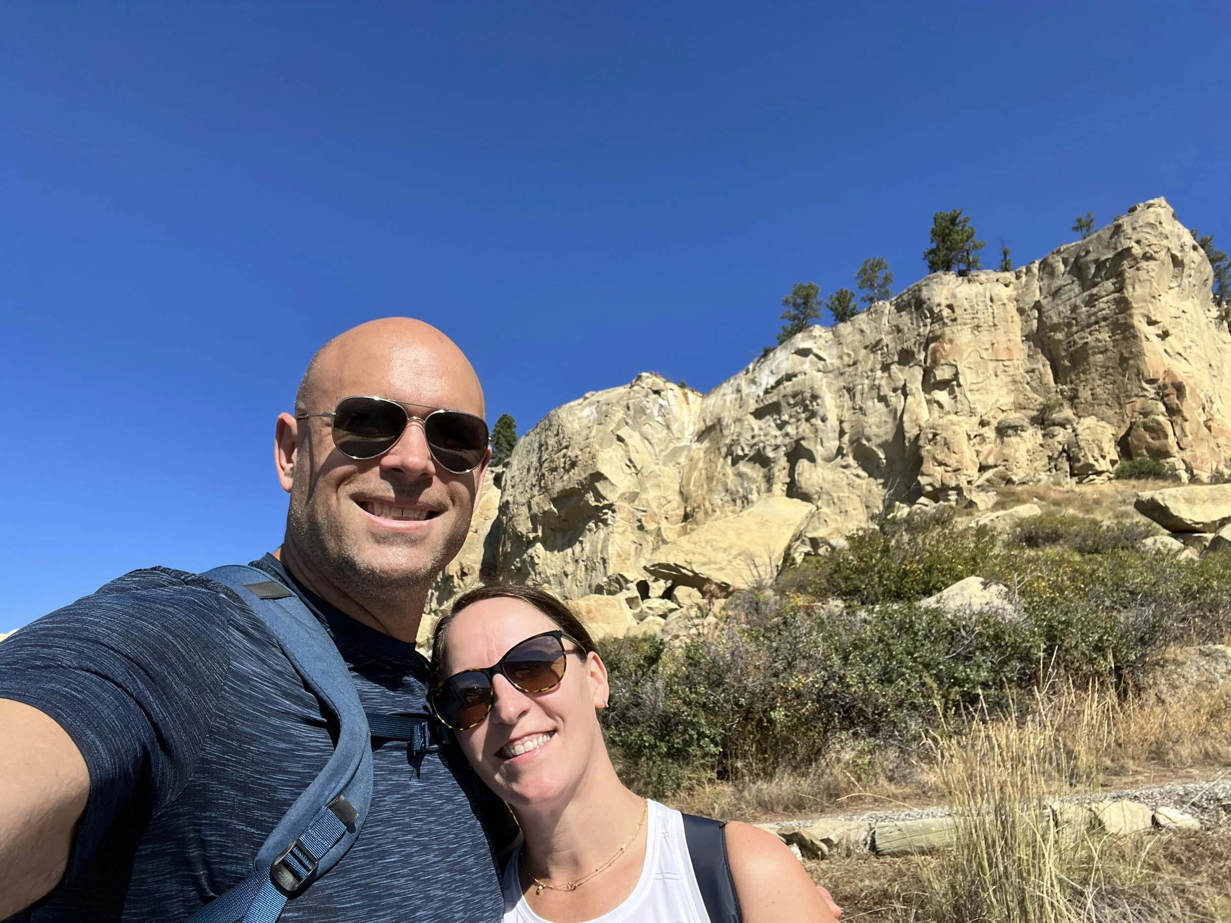 A group of three smiling people taking a selfie outdoors with rocky hills and trees in the background under a clear blue sky.