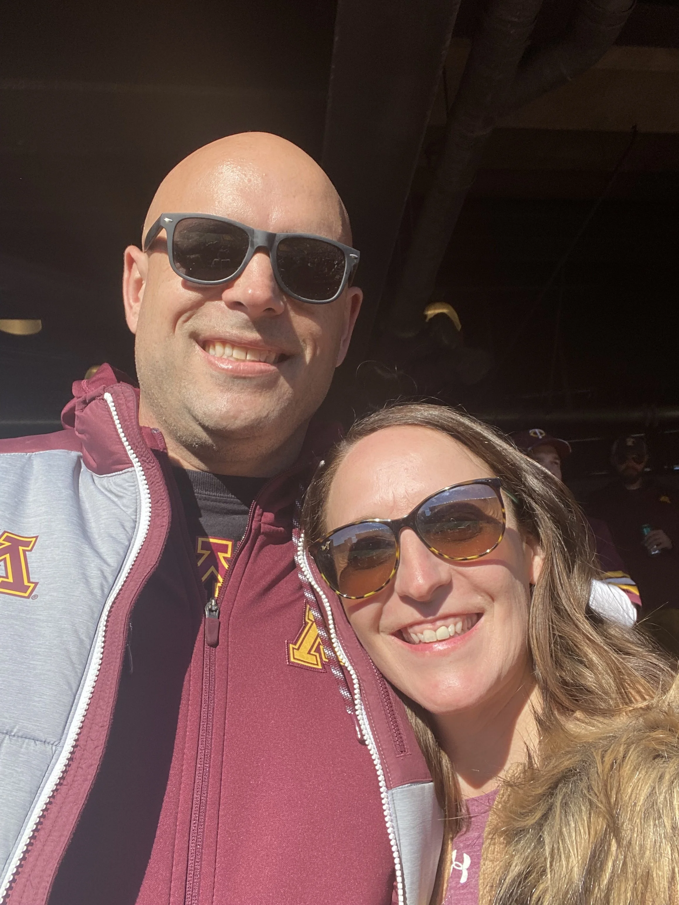 Close-up of a man and woman smiling, both wearing sunglasses, with people in the background.