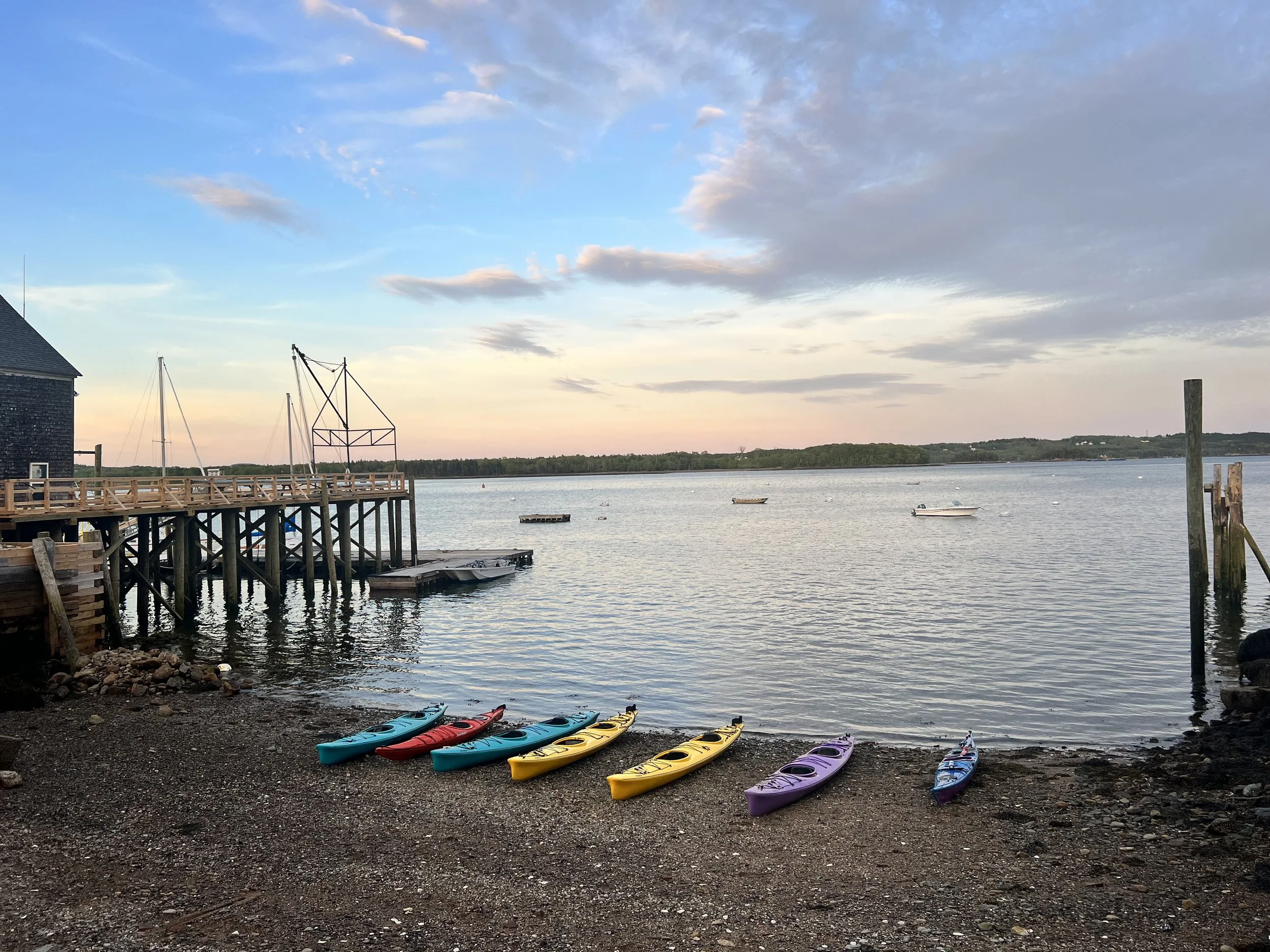 Colorful kayaks lined up on a shingle beach with a waterfront, small boats floating on the water, a wooden pier and house structure, and a partly cloudy sky at dusk or dawn.