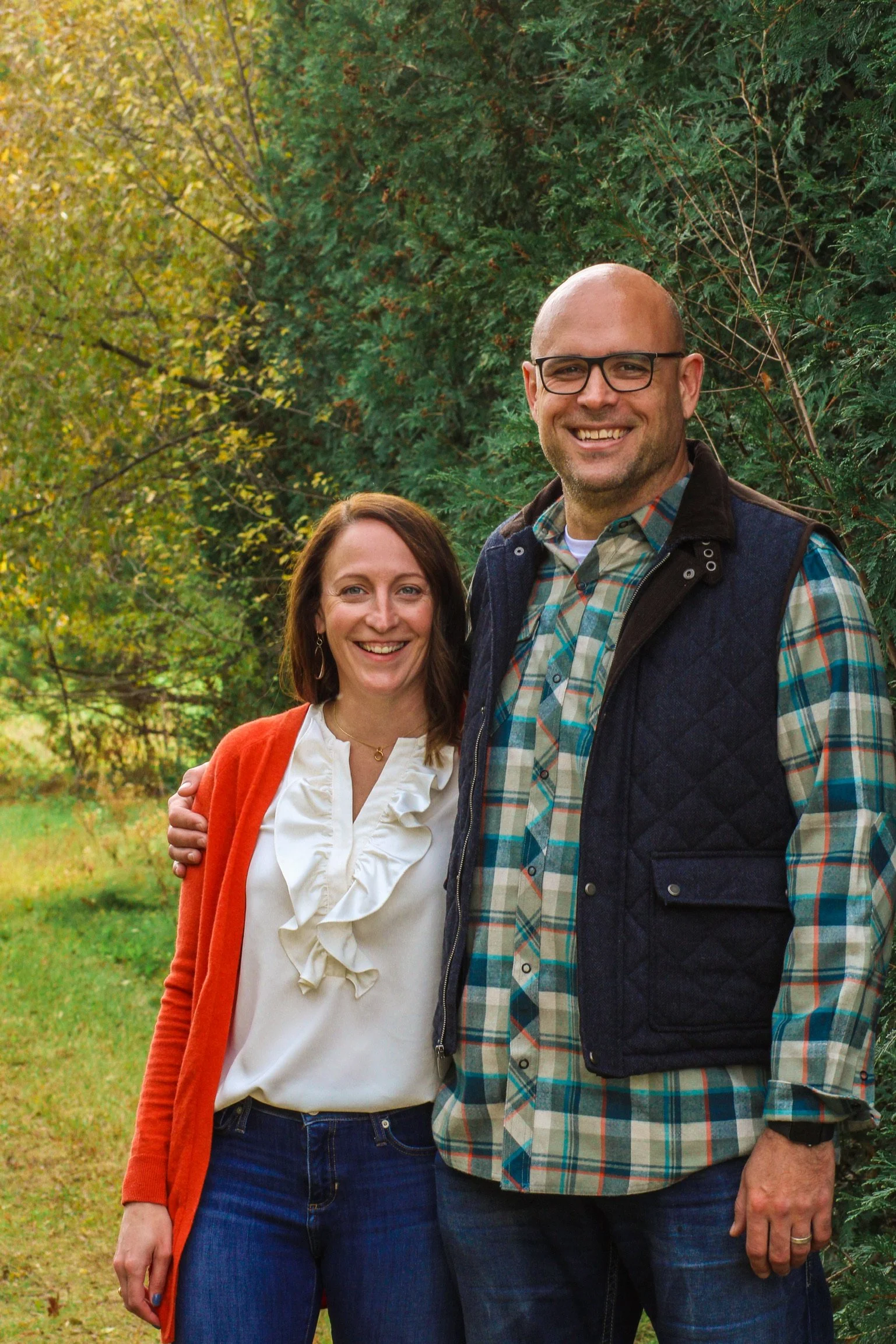 A smiling man with glasses, a beard, and a bald head stands next to a woman with brown hair, smiling, in an outdoor setting with green trees in the background. They are dressed casually, with the man wearing a plaid shirt and vest, and the woman wearing a white blouse and orange cardigan.