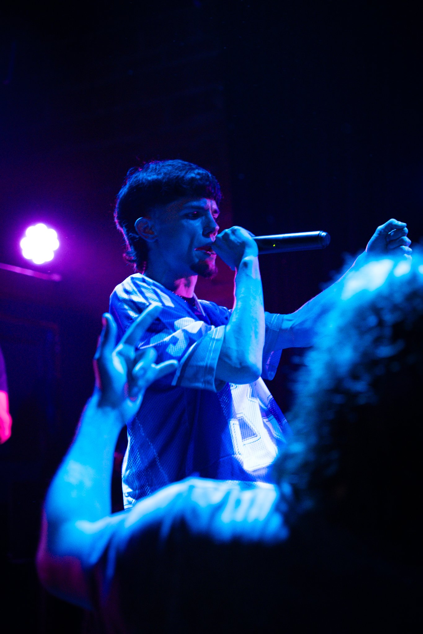 A male singer with short dark hair holding a microphone on stage, illuminated by blue and purple lighting, with hands of fans reaching out toward him.