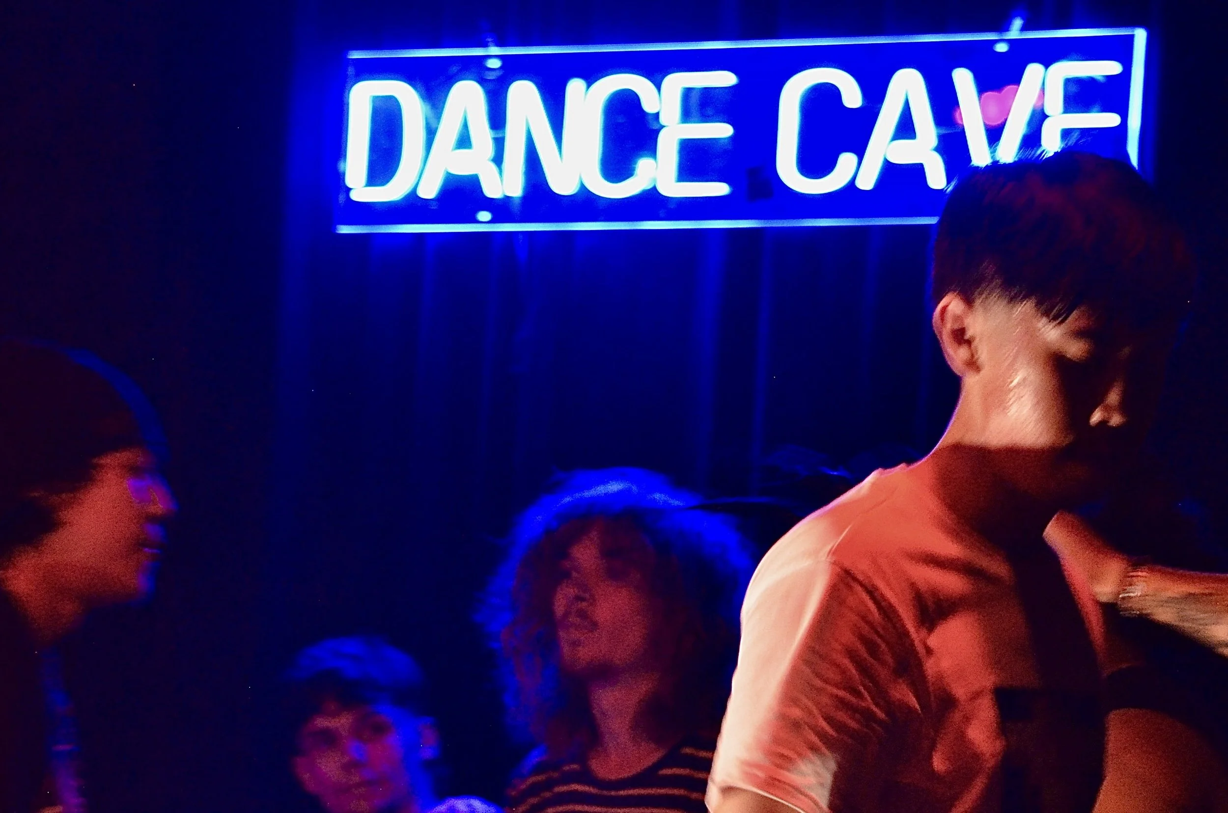 Bright blue neon sign that reads 'DANCE CAVE' in a dark room with several people dancing and socializing.
