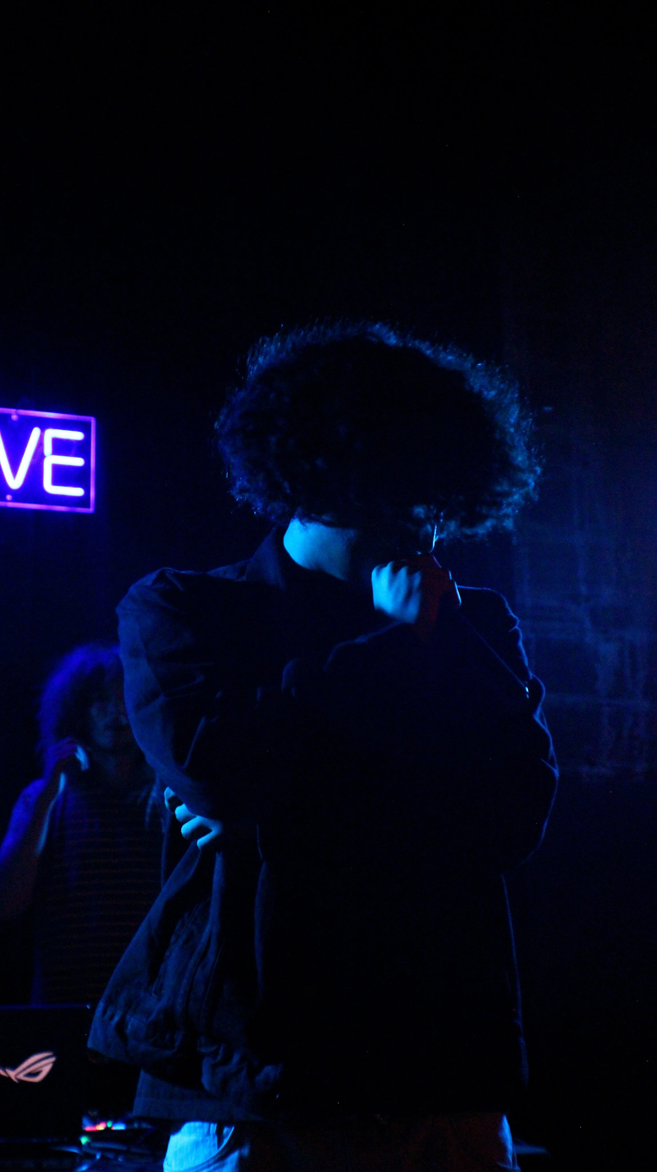 Person with curly hair wearing a dark jacket, holding a microphone, standing in a dark room illuminated by blue and purple neon lights; partial view of a neon sign reading 'LOVE' in the background; other individuals are visible in the background.