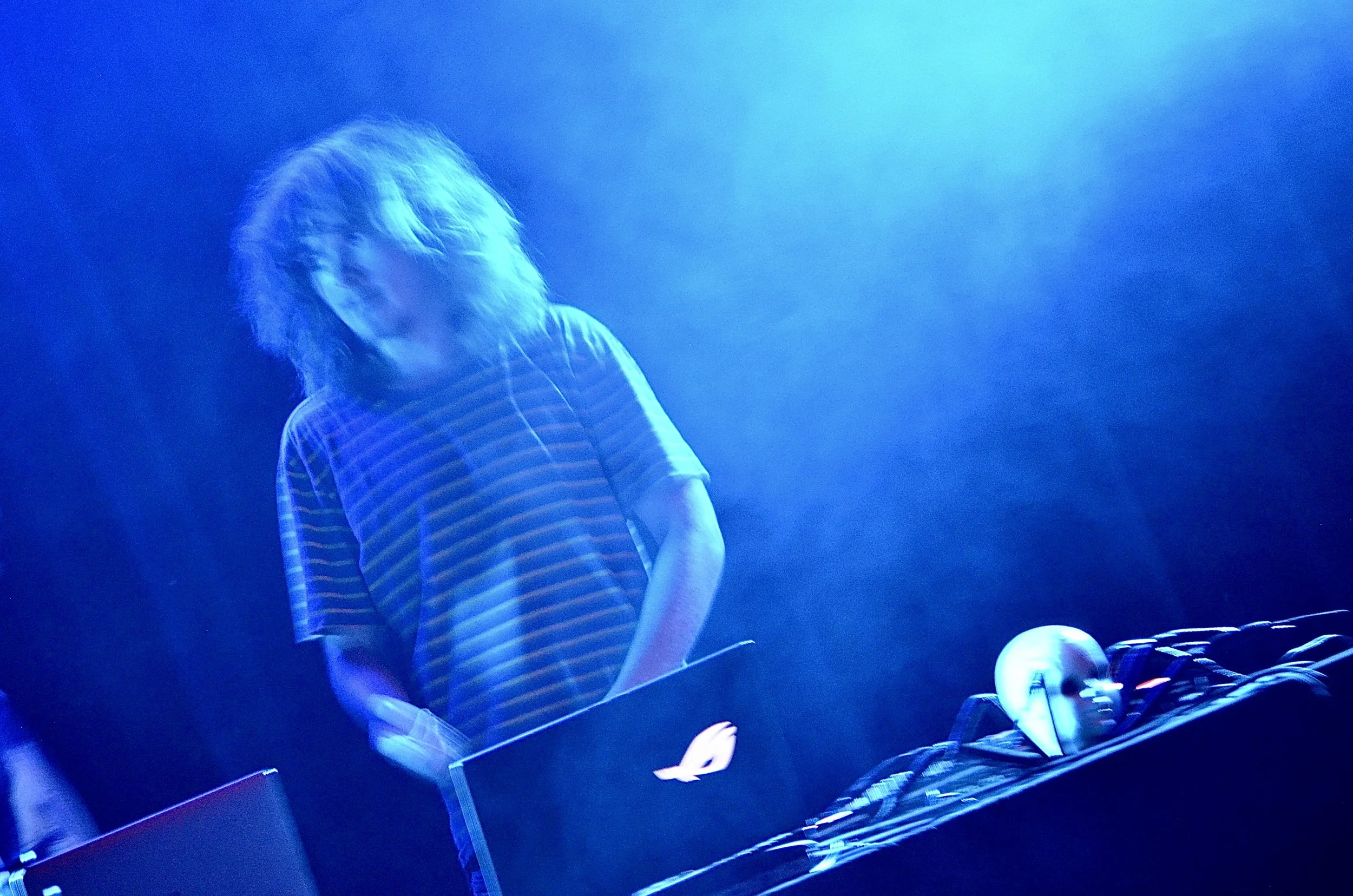 A person with light-colored hair wearing a striped shirt, standing at a DJ booth illuminated by blue lighting, with mixing equipment and a globe-shaped lamp nearby.