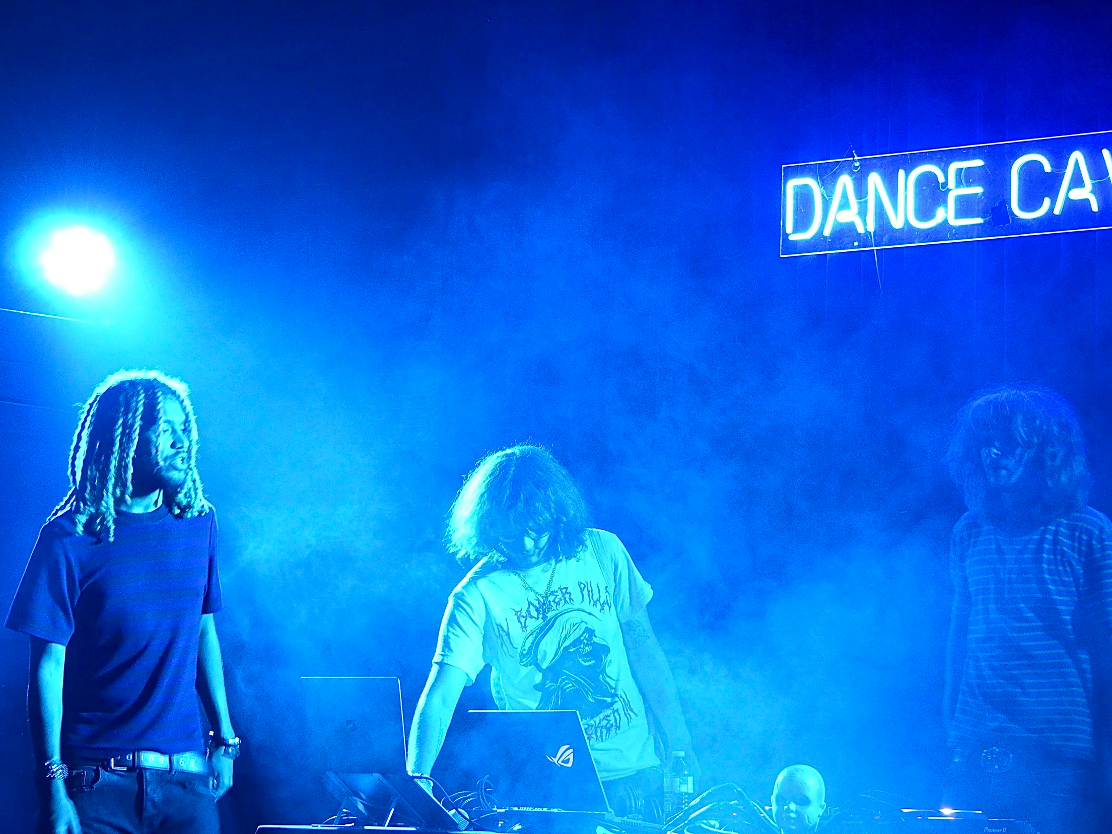 People standing in a dimly lit room with a bright blue neon sign that reads 'DANCE CAMP' on the wall.