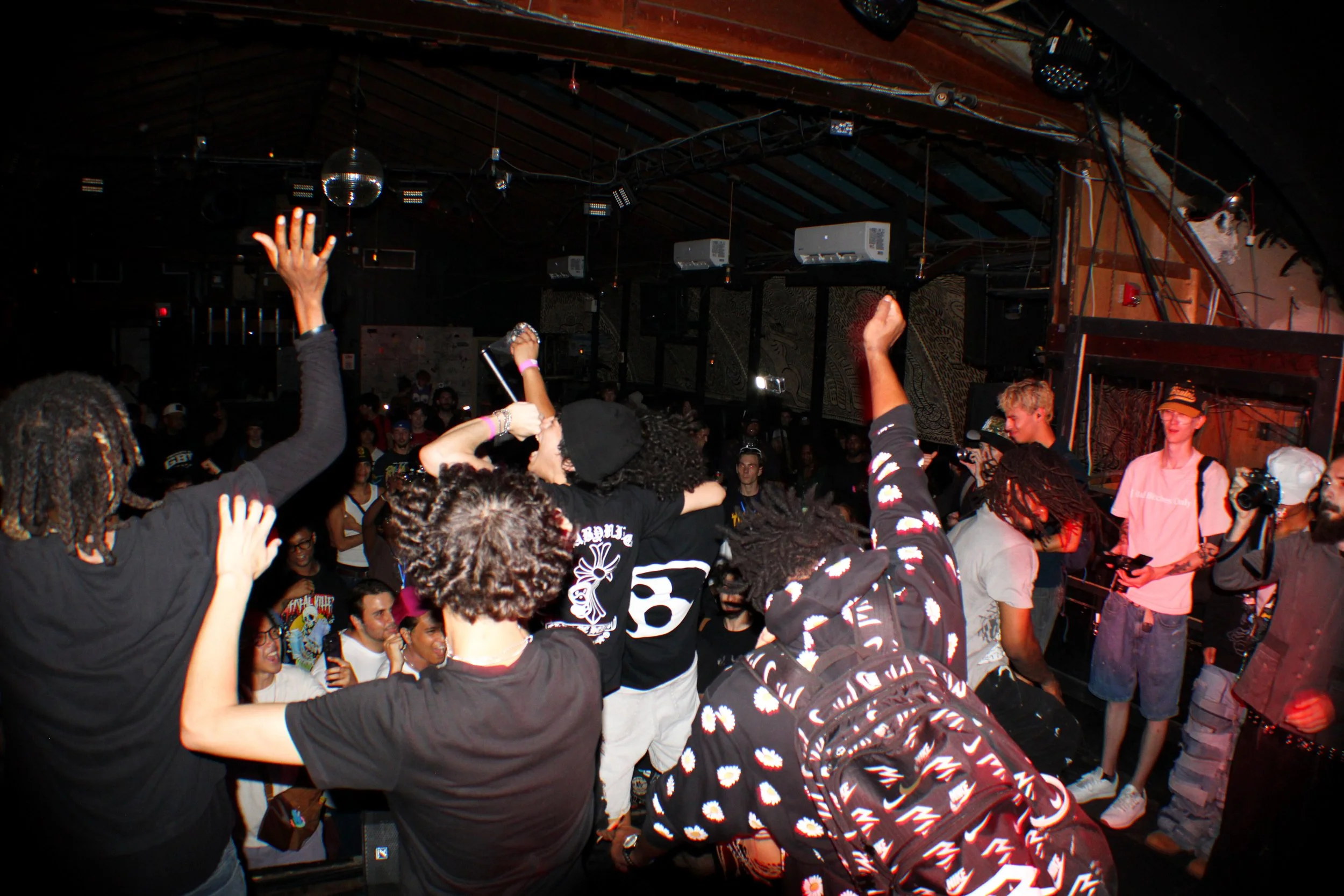 A crowded indoor concert or music event with people dancing and raising their hands, with stage lights, speakers, and a DJ or performer in the background.