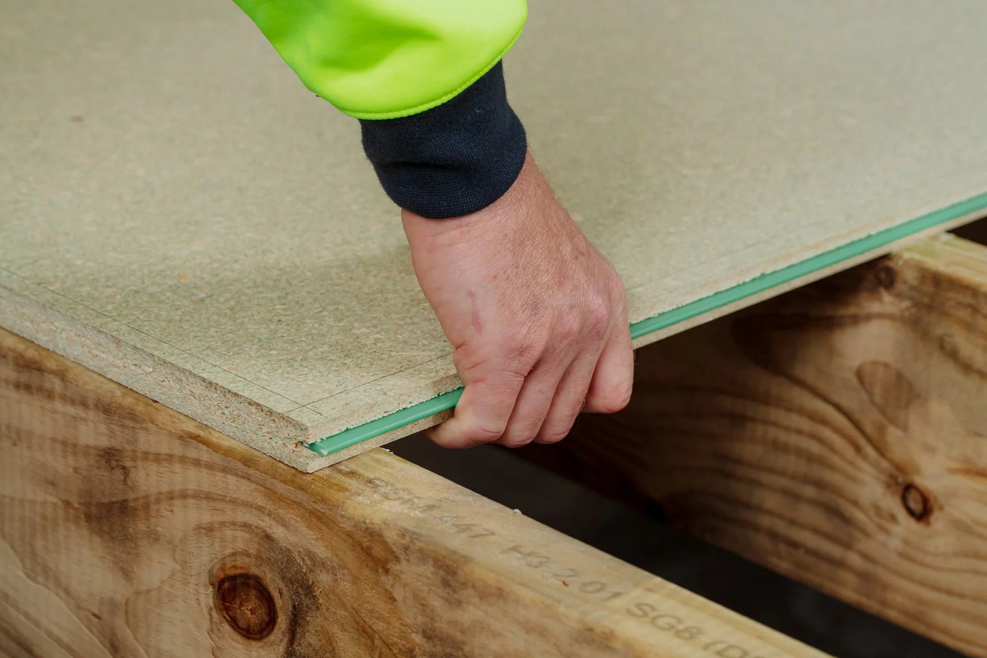 A person measuring a piece of particle board with a pencil, marking guide lines on the board, while working on a construction project. The person's hand is visible, wearing a black sleeve, with a green and black shirt or jacket.