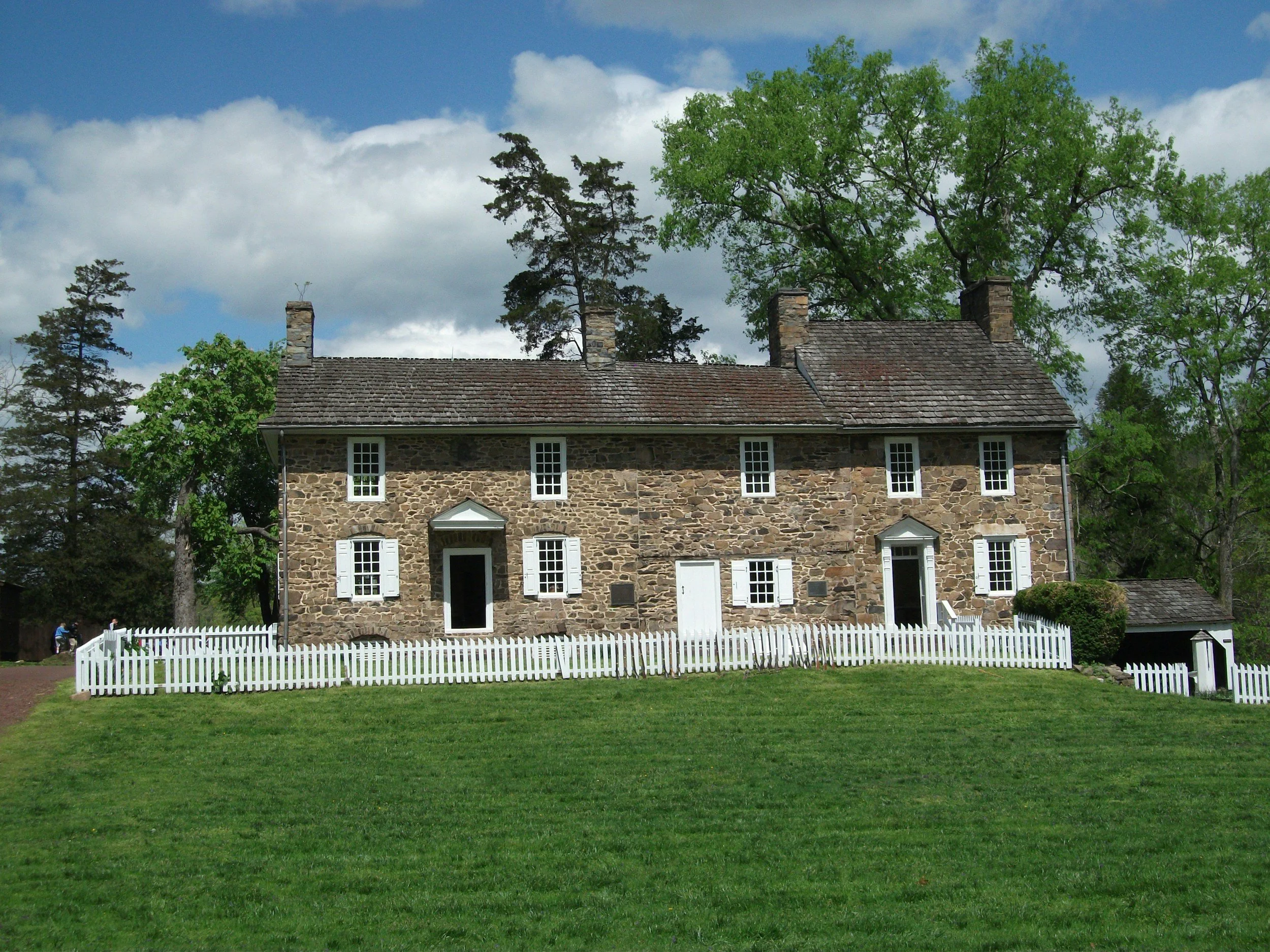 A stone house with multiple windows and a door, surrounded by a white picket fence, green grass, and tall trees under a partly cloudy sky.