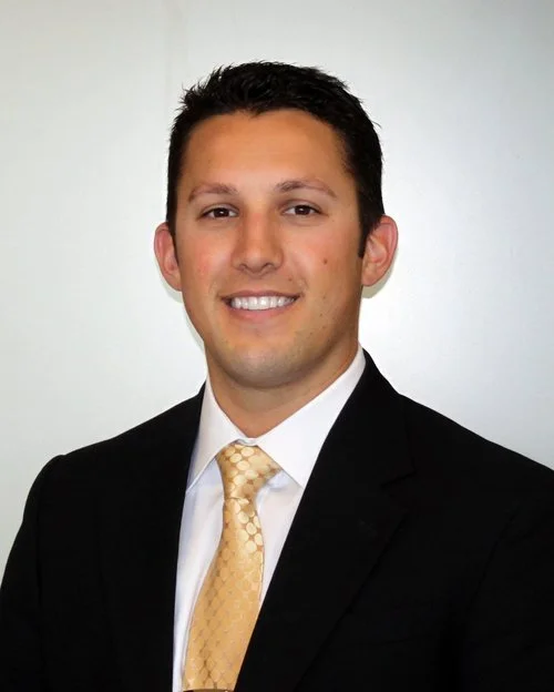A young man in a black suit with a white shirt and yellow tie, smiling against a plain white background.