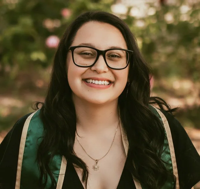 A young woman wearing glasses, smiling, dressed in graduation attire outdoors.
