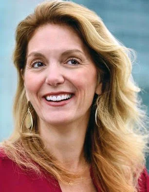 A smiling woman with wavy blonde hair, wearing a maroon top and earrings, standing outdoors against a blue background.