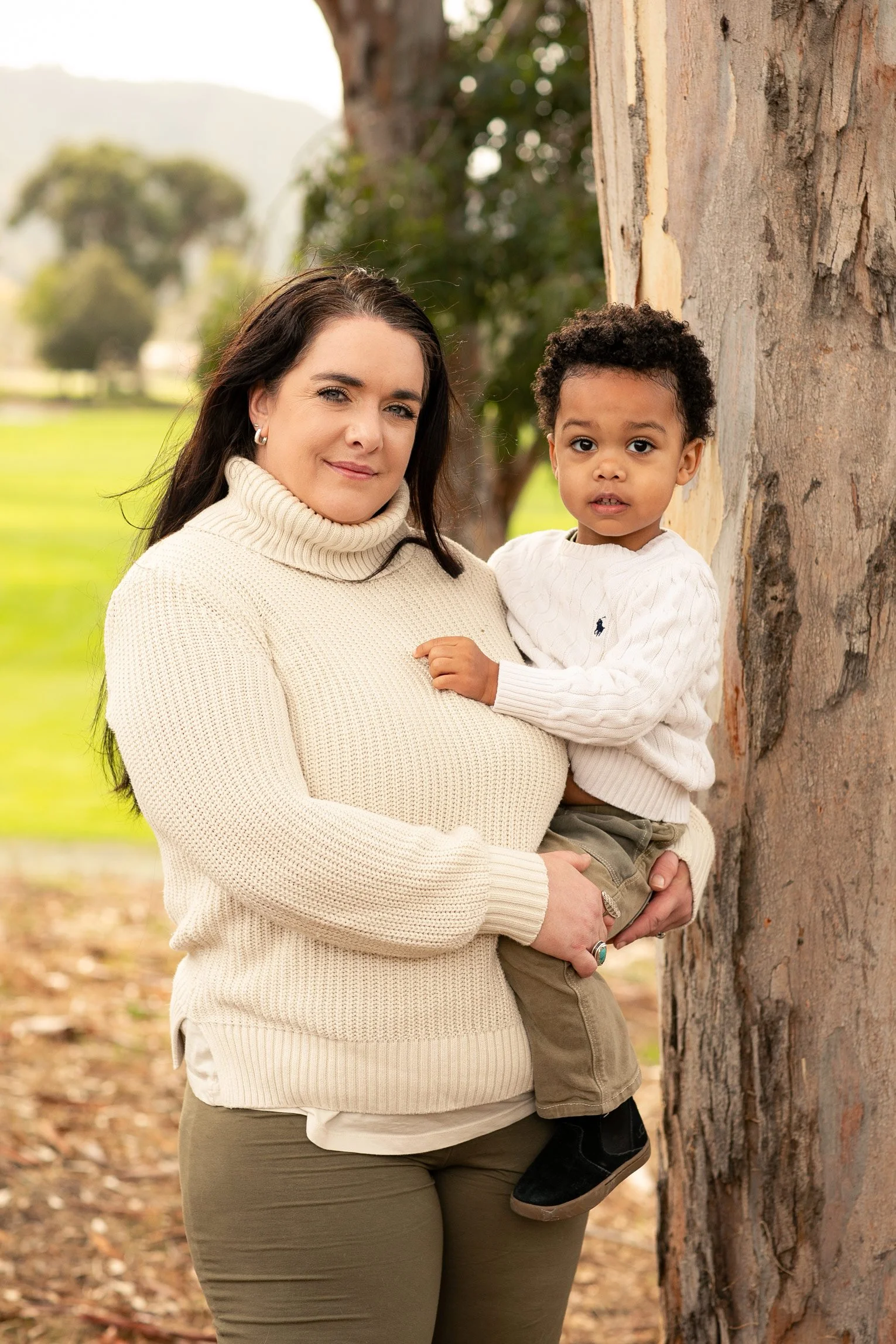 A woman with dark hair in a beige turtleneck sweater holding a young boy with black curly hair in a white sweater and khaki pants, standing outdoors next to a large tree in a park.