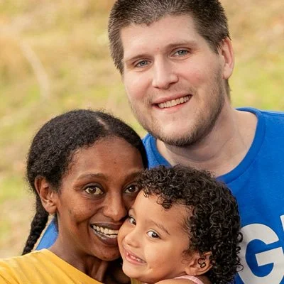 A smiling family of three outdoors, with a woman, a man, and a young child, all close together.