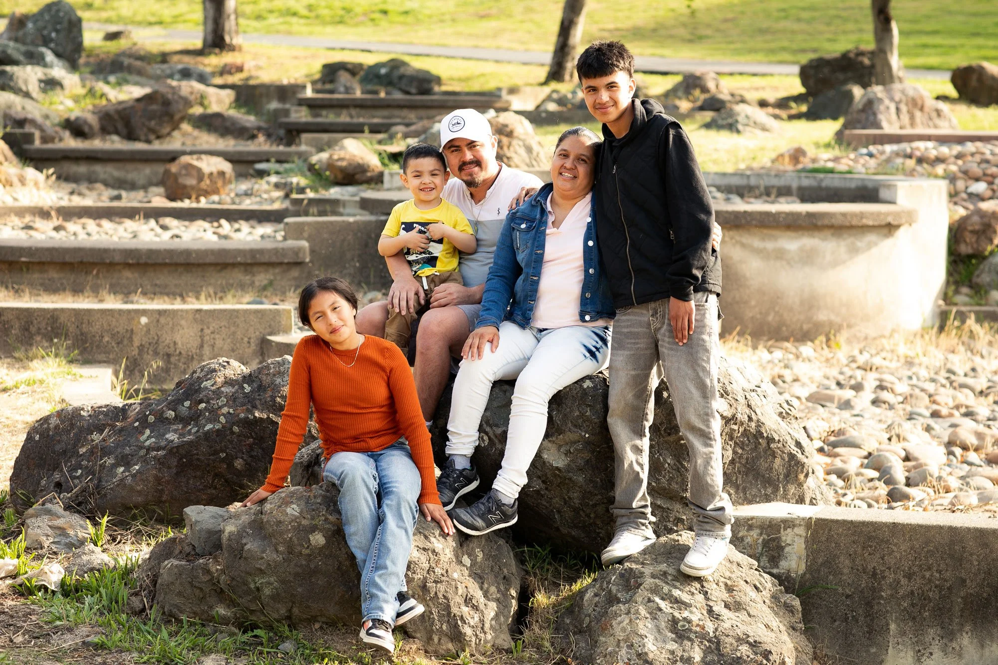 A multigenerational family of six people sitting and standing on rocks outdoors, smiling at the camera, with a rocky and grassy park area in the background.