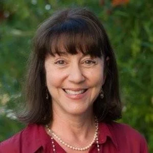 Portrait of a smiling woman with dark brown hair and bangs, wearing a maroon top, pearl necklace, and earrings, outdoors with green foliage background.