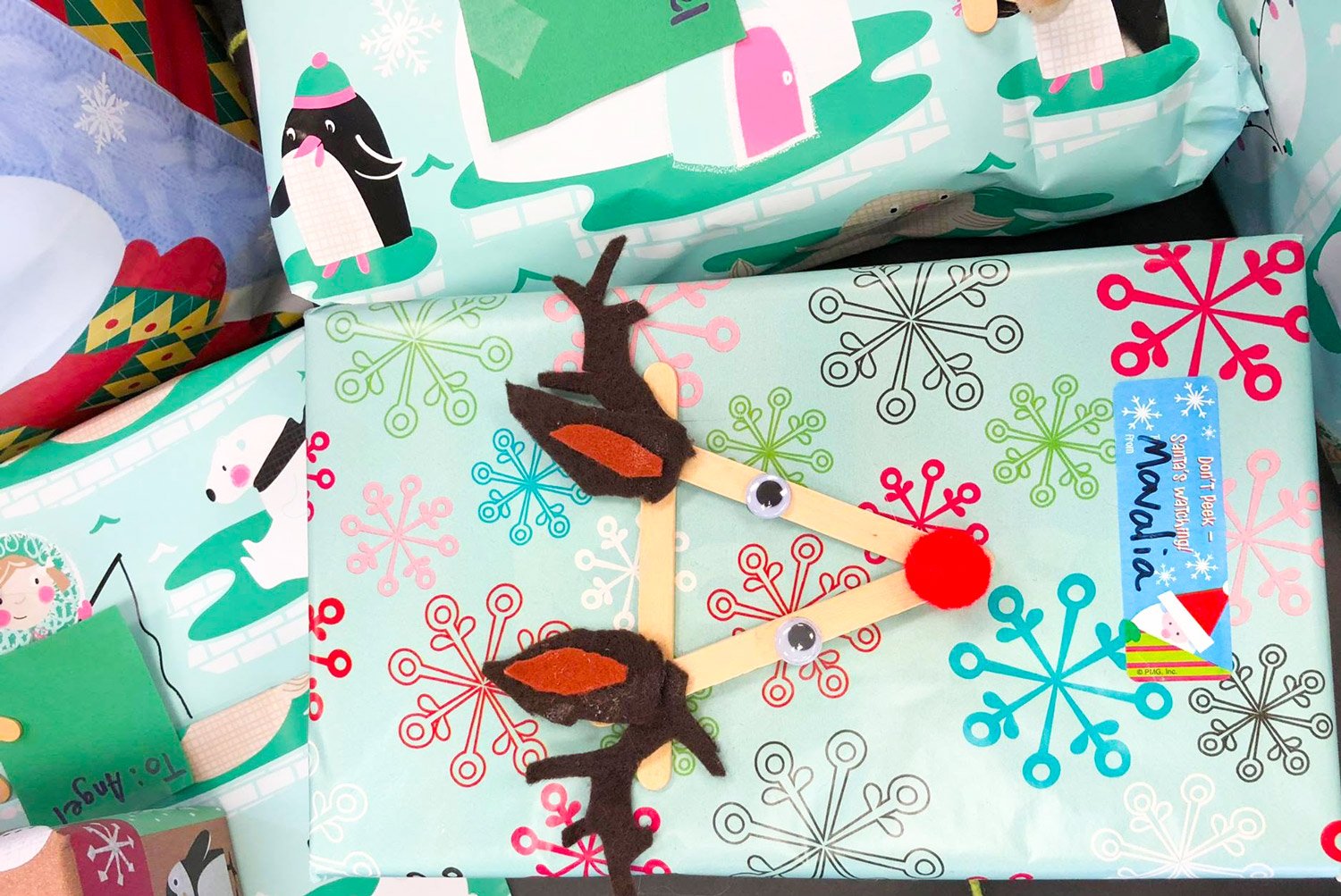 A handmade craft resembling a reindeer face, made with popsicle sticks, felt, googly eyes, and a red pom-pom, placed on a Christmas-themed gift bag decorated with snowflakes.