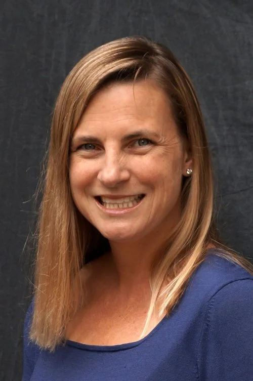 A woman with shoulder-length light brown hair, wearing a blue top, smiling at the camera against a dark background.