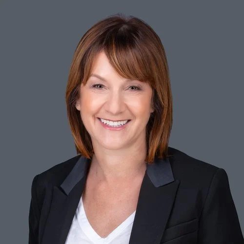 Portrait of a smiling woman with shoulder-length brown hair wearing a black blazer and white shirt against a gray background.