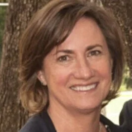 A woman with short brown hair smiling outdoors in front of a tree.