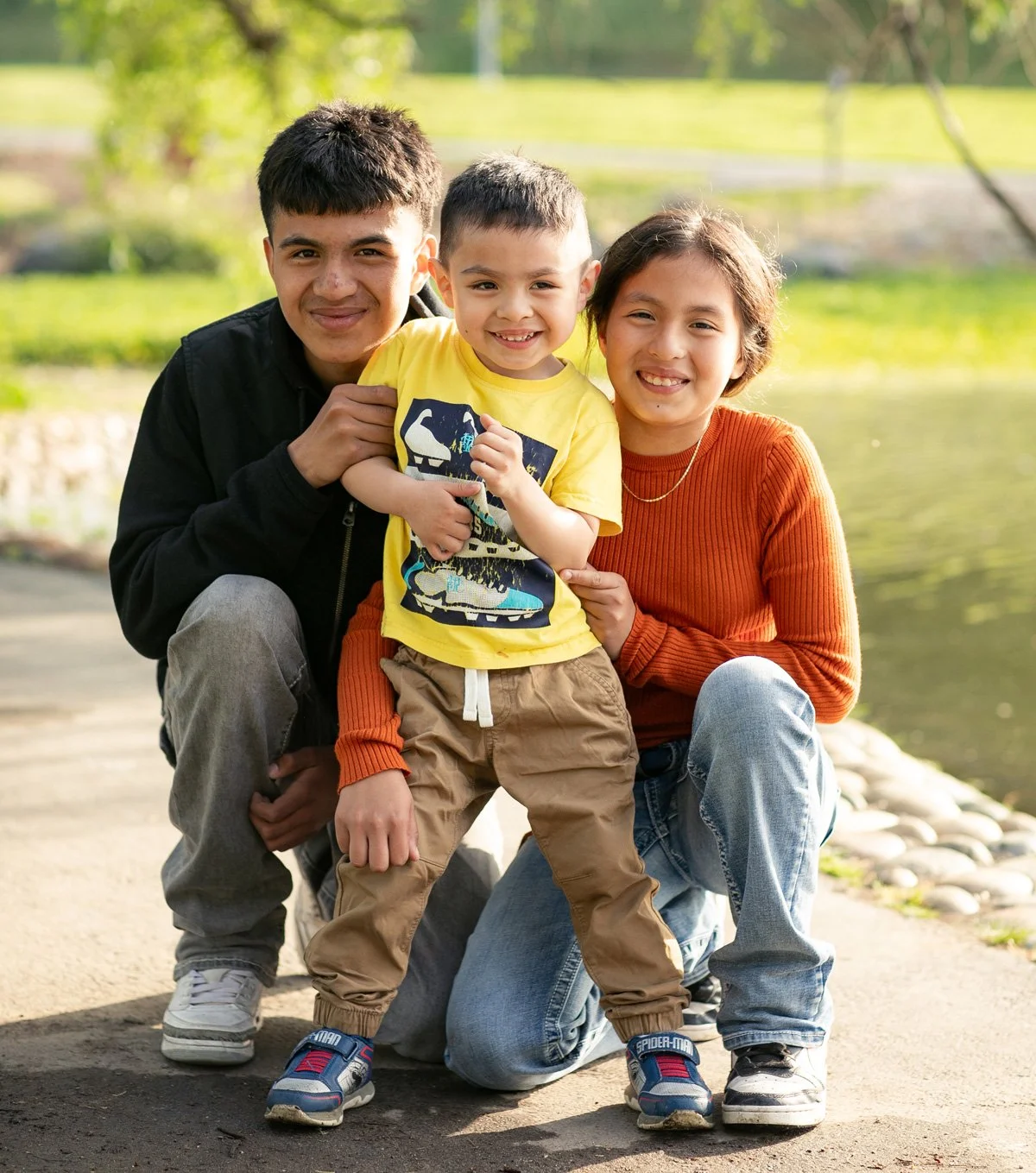 A family of three, including a teenage boy, a young boy, and a woman, smiling and crouching together outdoors near a pond or river with greenery in the background.